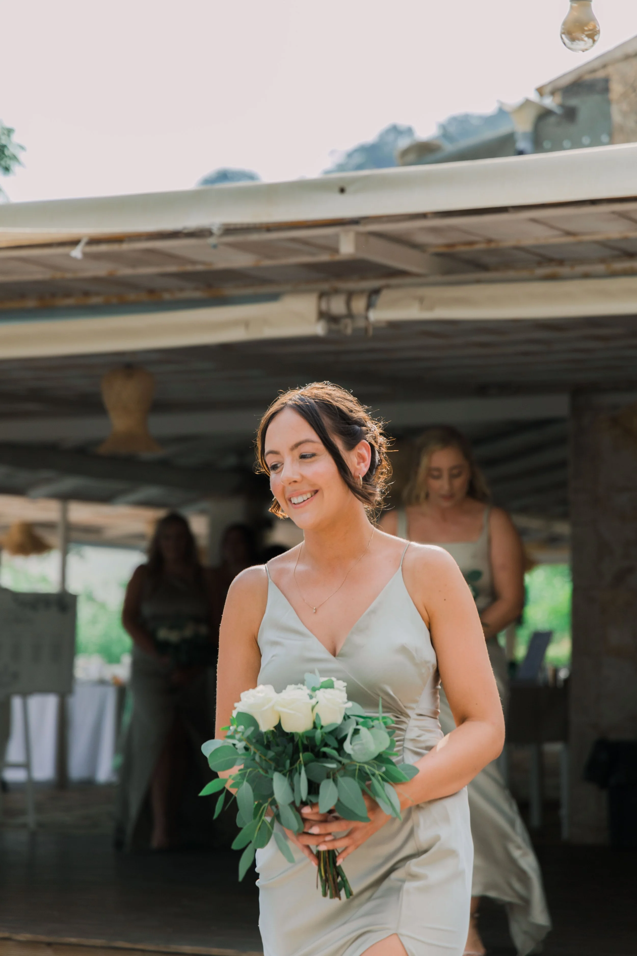 A woman in a light-colored satin dress holding a bouquet of white roses and greenery, smiling at an outdoor event, with other women in the background.