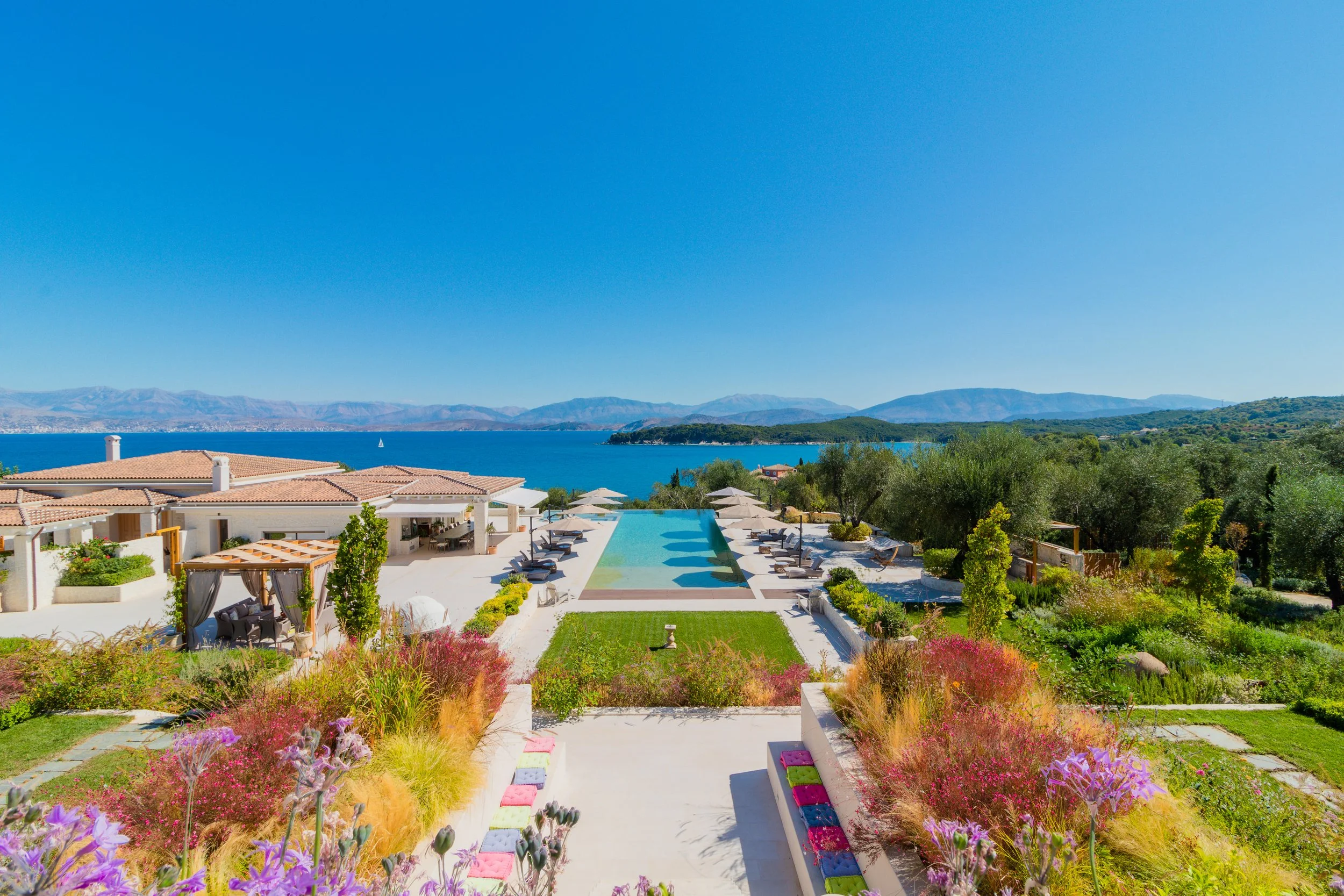 Luxury resort with a swimming pool overlooking a lake, surrounded by greenery and mountains under a clear blue sky.