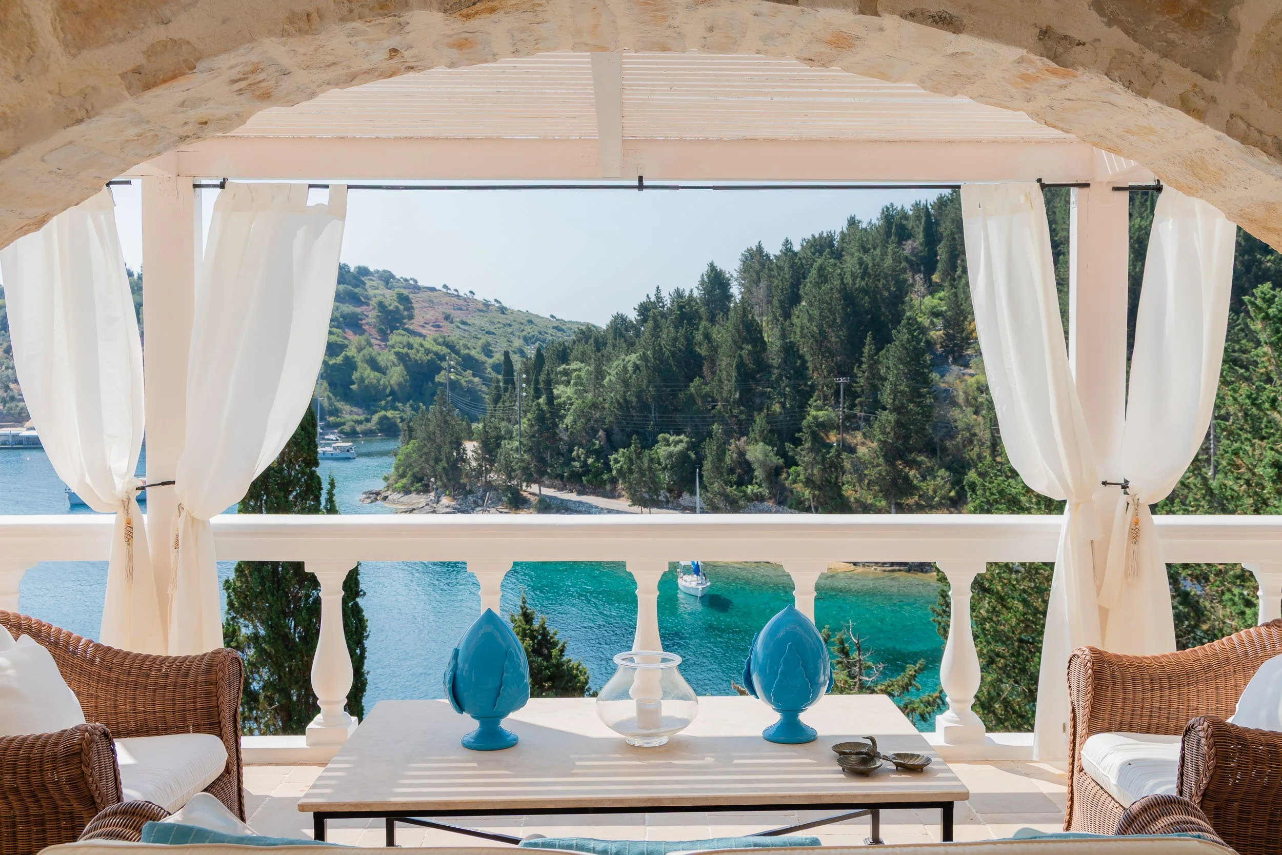 Terrace with wicker chairs and a table, overlooking a bay with trees and a sailboat, framed by white curtains