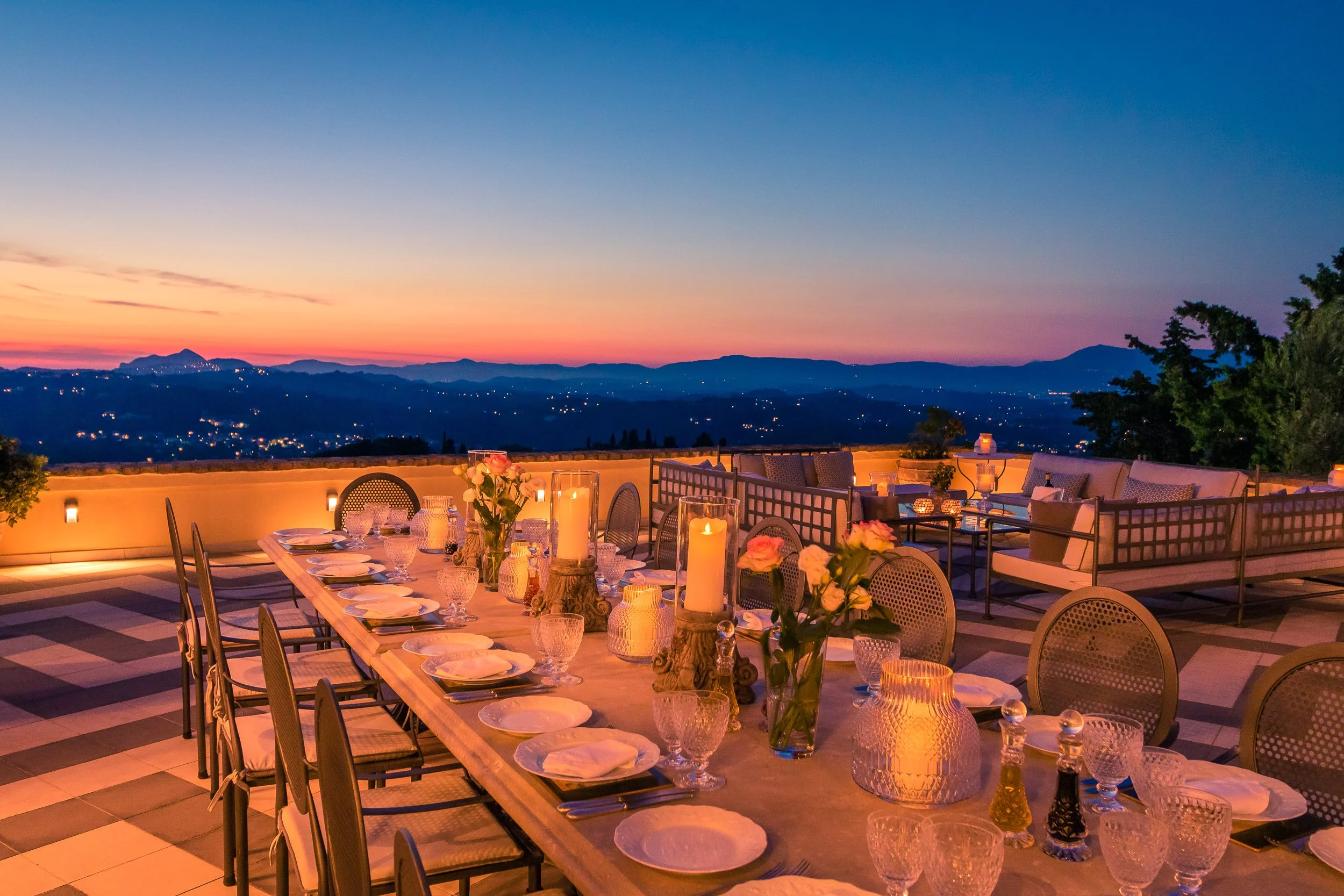 Elegant outdoor dining table set for a meal with candles, flowers, and glassware, overlooking a mountain range at sunset.