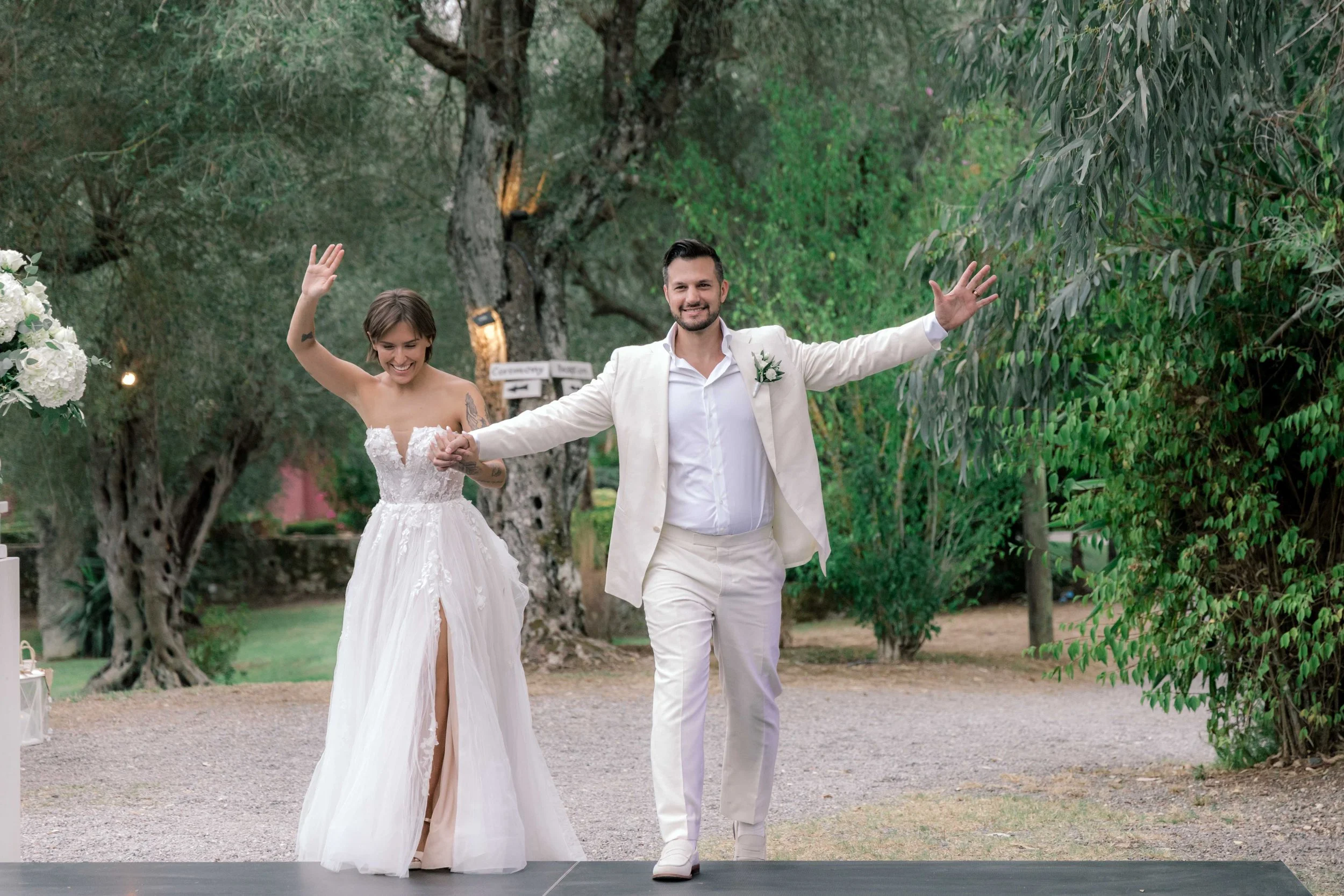A newlywed couple walking outdoors on a gravel pathway surrounded by trees and greenery, holding hands and smiling, with the groom wearing a cream suit and the bride in a white wedding dress.