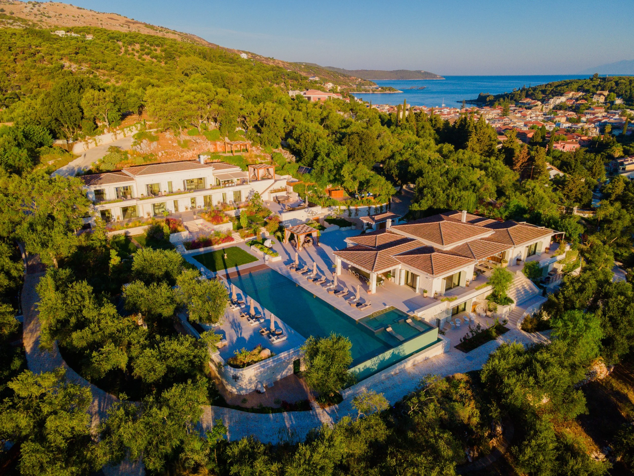 An aerial view of a luxurious seaside estate with several buildings, a large swimming pool, and surrounding lush greenery near the coast with hills and a small town in the background.