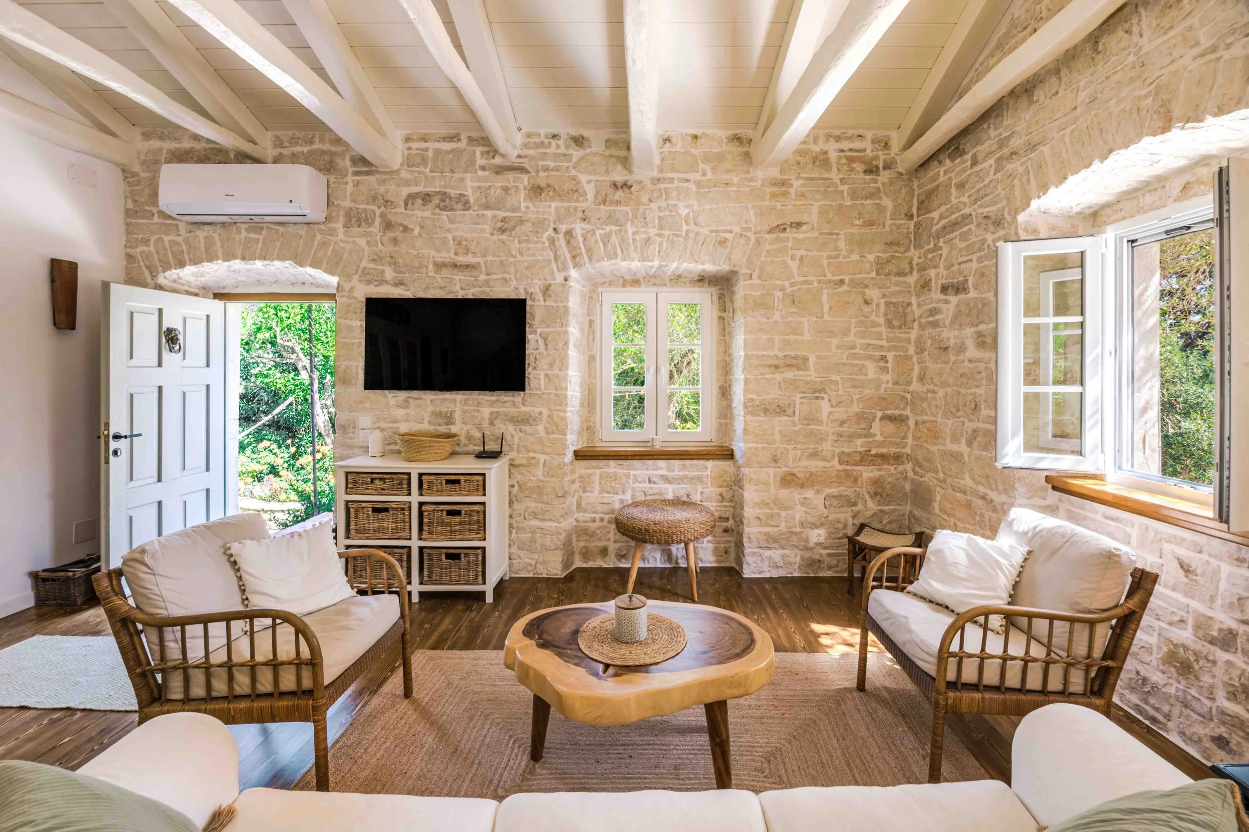 Living room with stone walls, wooden beams ceiling, open windows with greenery outside, white sofa, rattan chairs, wooden coffee table, TV on wall, and wicker furniture.