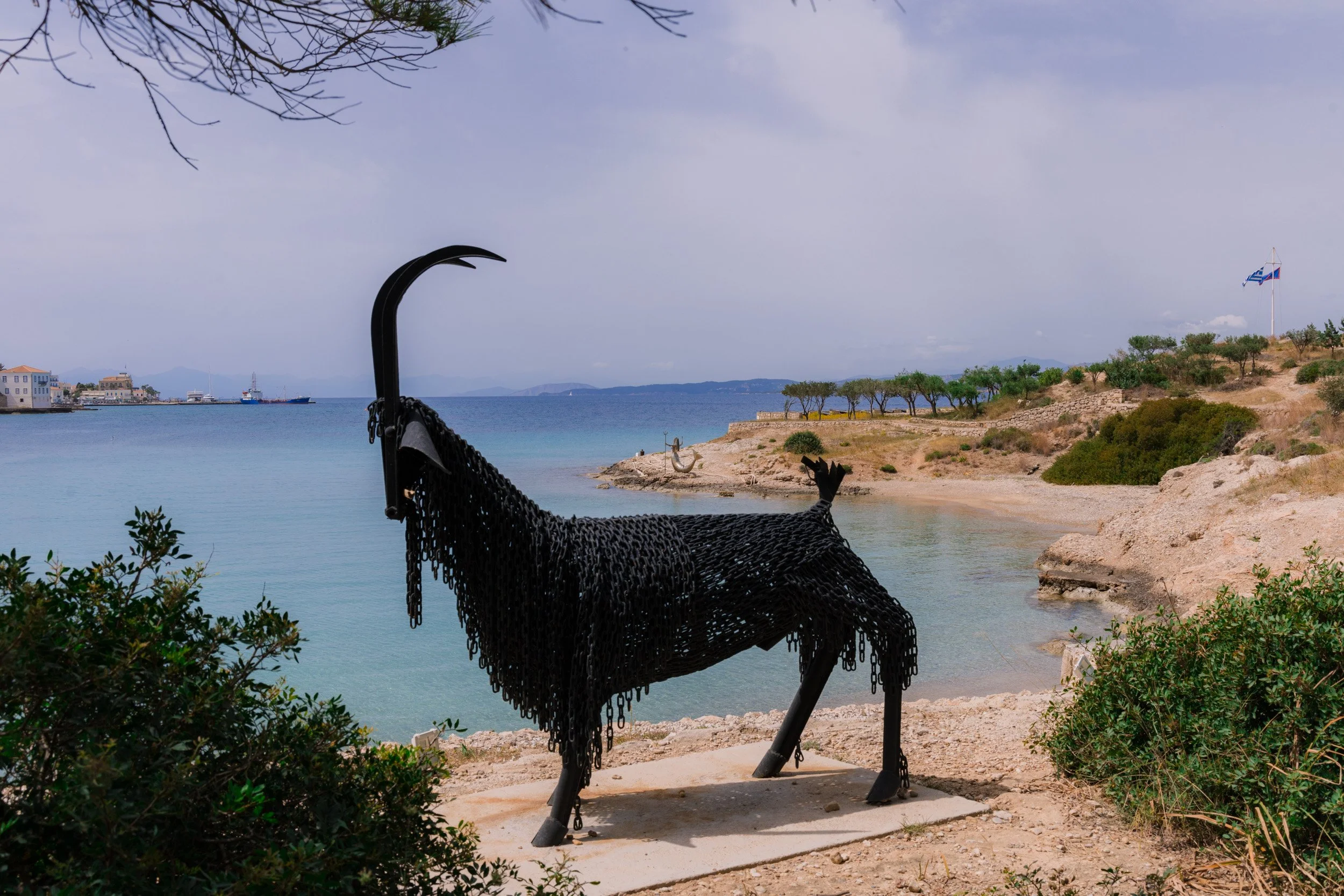 A metal sculpture of a goat, made of black chains, standing on a concrete base near the shore, overlooking a calm sea with a coastal landscape, trees, and a Greek flag in the background.