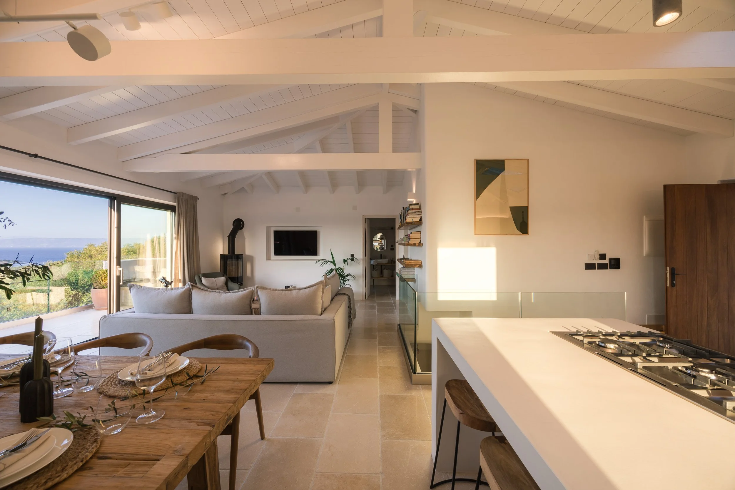 Open-concept living room and kitchen with white beams ceiling, sliding glass door, dining table with dishware, beige sofa, potted plants, wall-mounted TV, bookshelf, and a view of the outdoors.