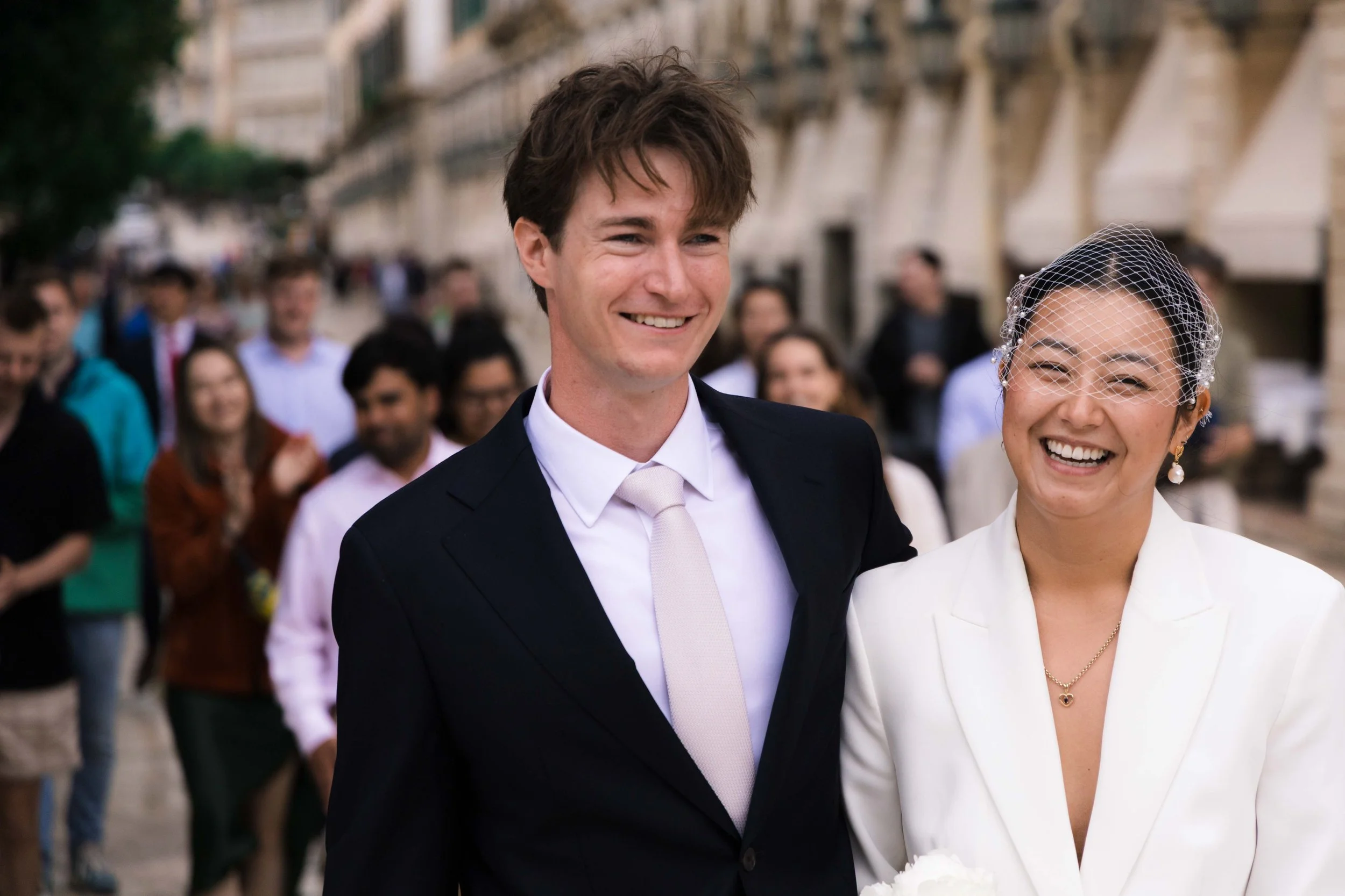 A man in a dark suit and a woman in a white suit smiling at a wedding celebration with friends in the background.