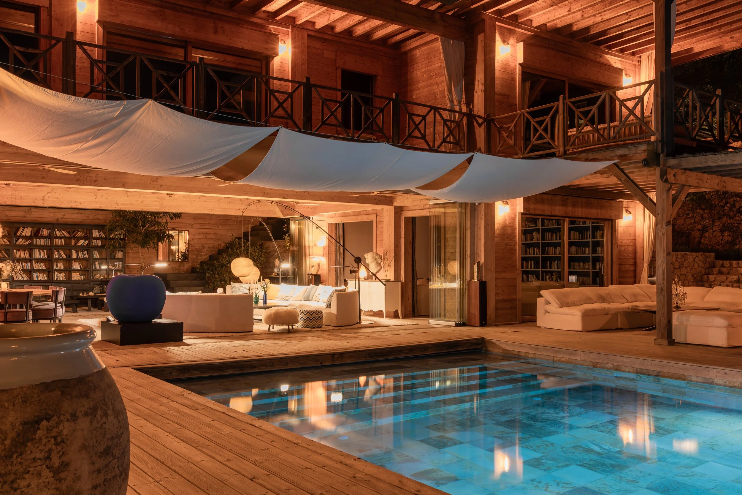 Luxury wooden house with a swimming pool, cozy outdoor lounge area, bookshelves, sofas, and warm lighting at night.
