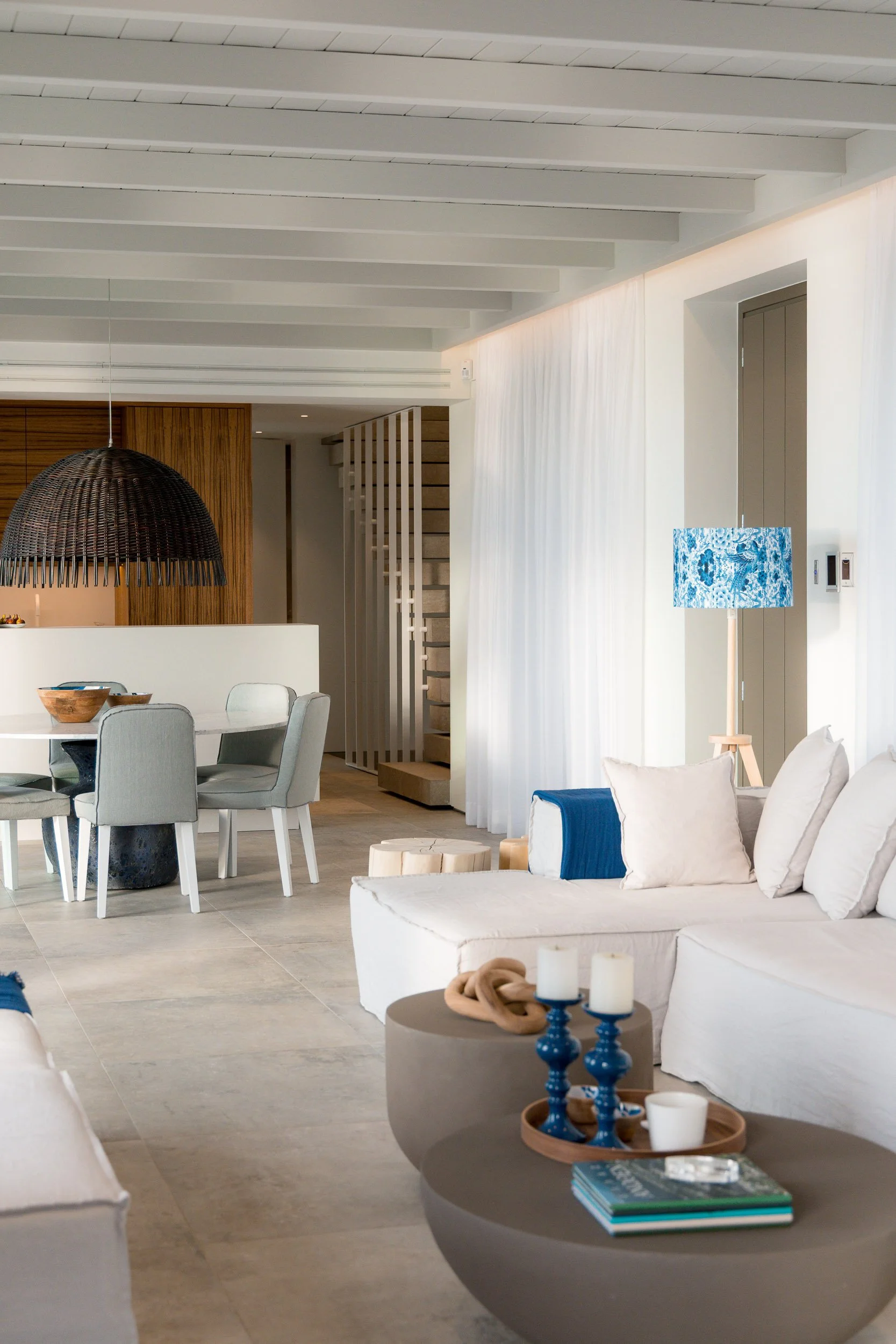 Modern living room with white sofa, blue accents, and contemporary decor, open to dining area with round table and gray chairs, and a staircase in the background.
