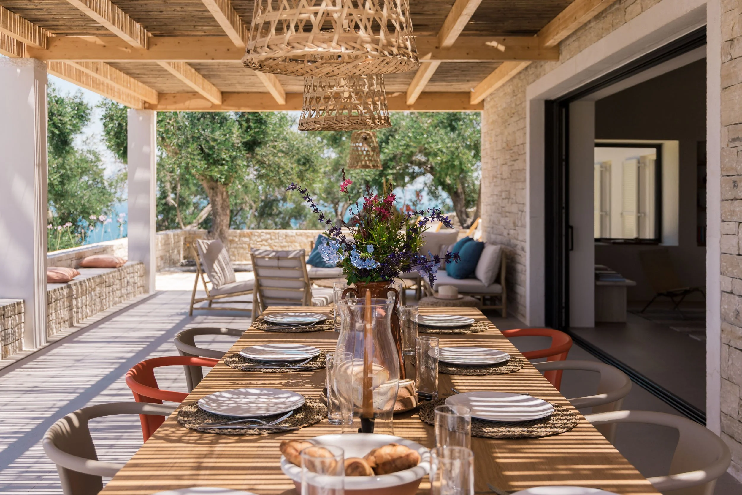 Outdoor dining area with a long wooden table set for a meal, featuring plates, glasses, and a central flower arrangement, under a wooden pergola with woven pendant lights, with trees visible in the background.