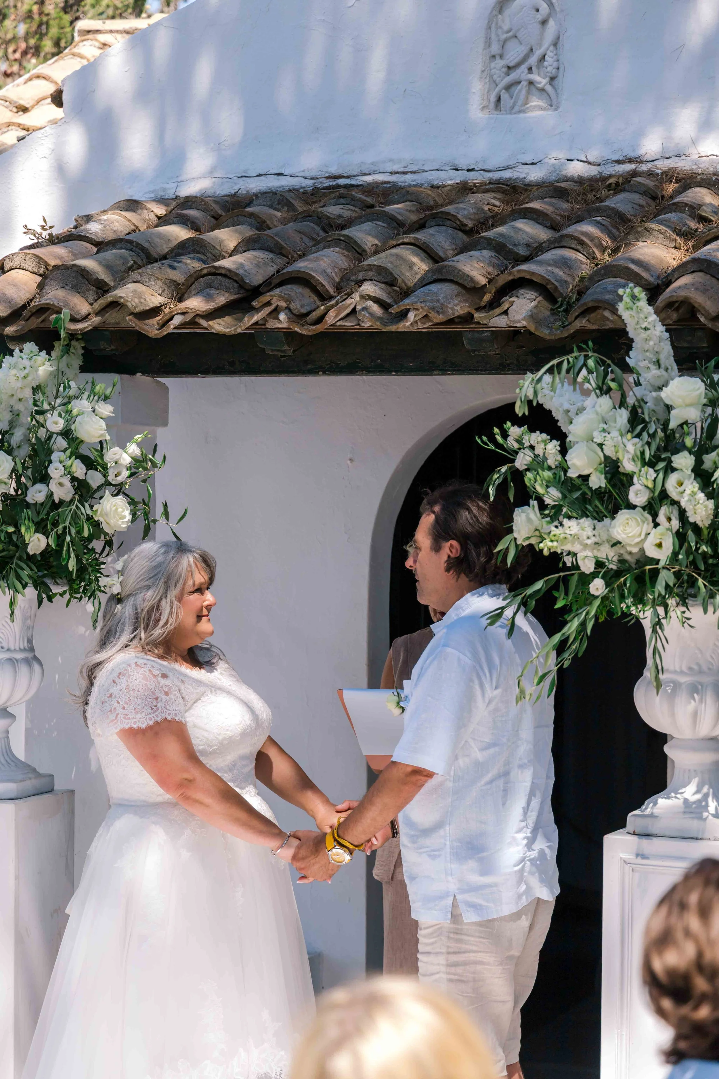 A wedding ceremony with a bride and groom holding hands, standing in front of a white wall with a tiled roof, decorated with large white flower arrangements on either side.