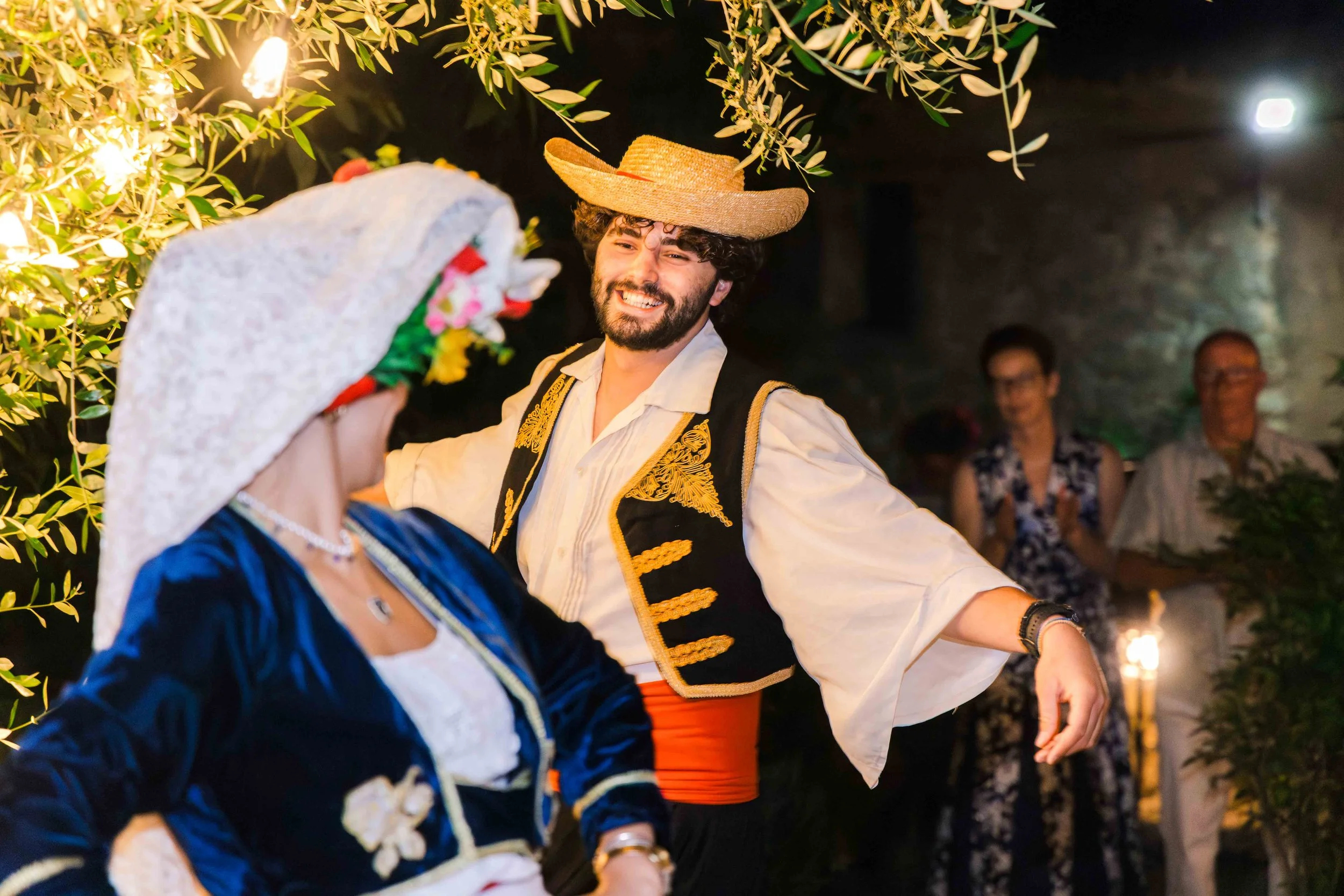 People at a lively nighttime celebration, with a man wearing a straw hat and traditional Mexican attire dancing and smiling, while a woman in a colorful dress and wide-brimmed hat looks on. The scene is decorated with string lights and greenery.