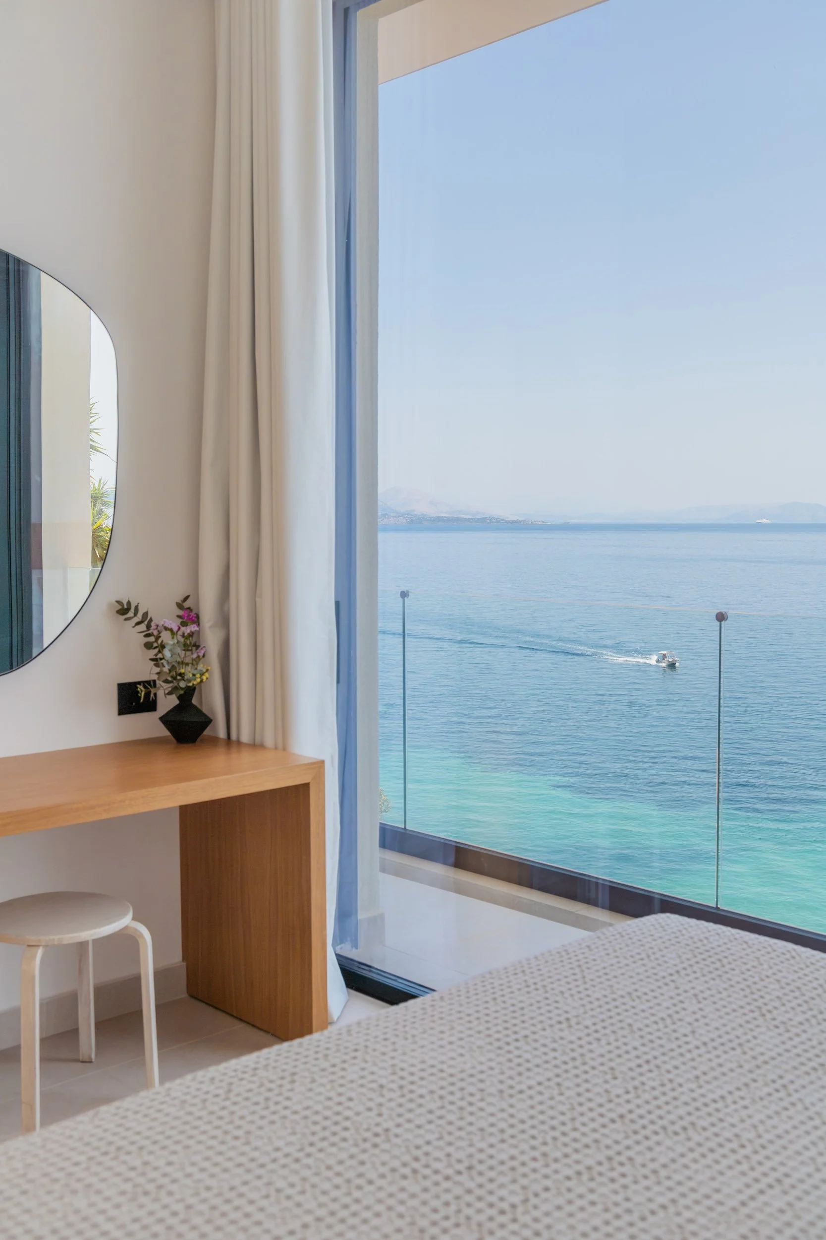 A hotel room with a glass door balcony overlooking a blue ocean, a boat on the water, white curtains, a wooden desk with a small black vase and pink flowers, a round mirror on the wall, a white stool, and part of a bed with white sheets.
