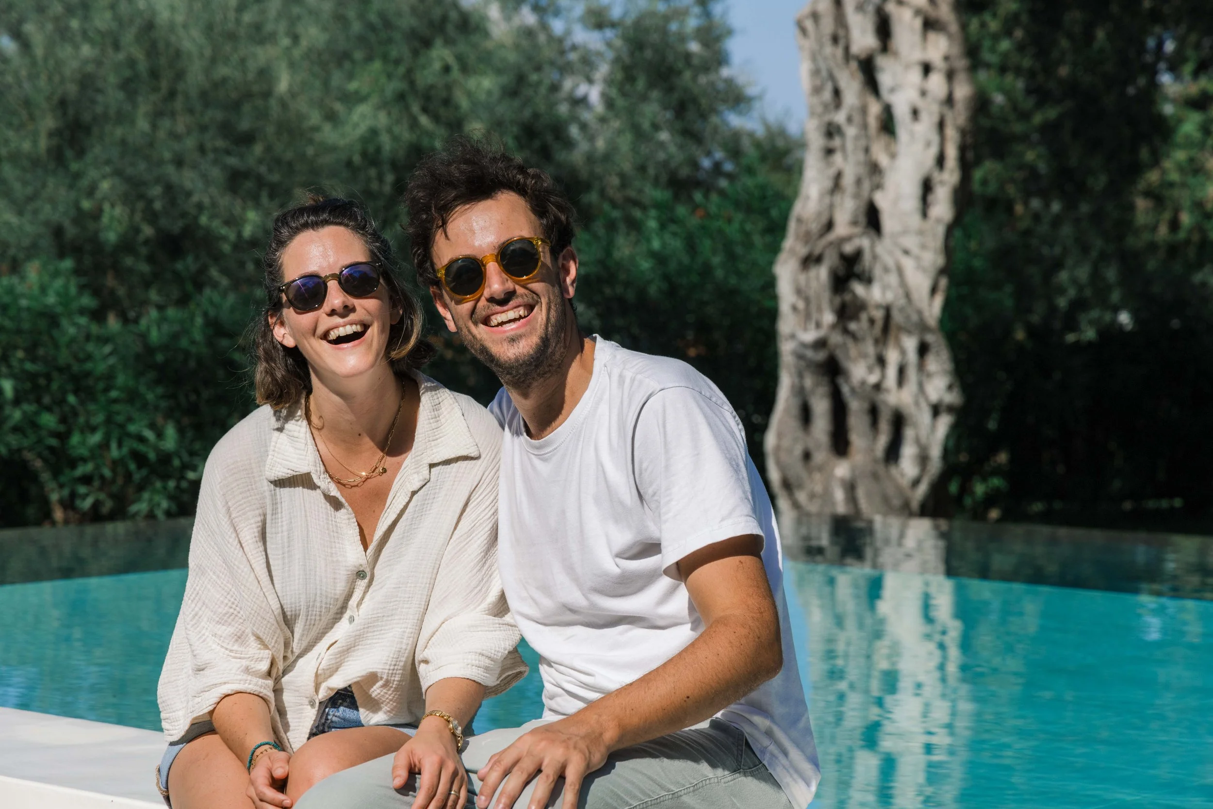 A smiling couple sitting by a swimming pool with trees in the background, enjoying a sunny day.