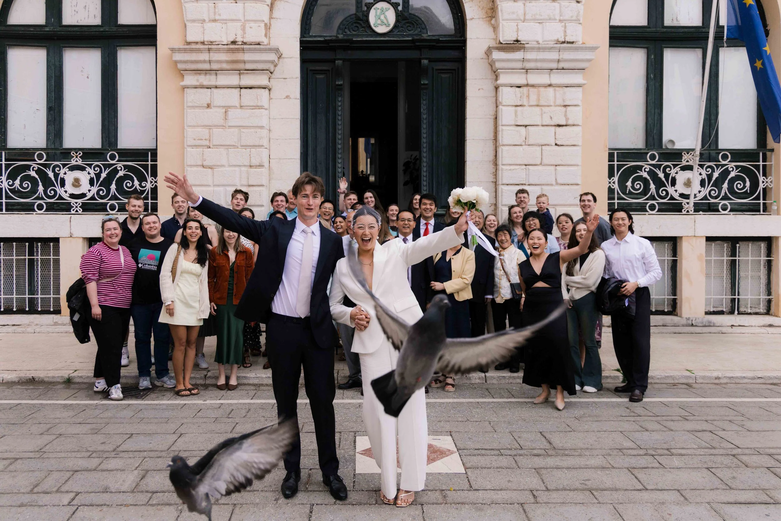 A group of people celebrating a wedding in front of a building, with two people in the center holding hands and a dove flying in front.