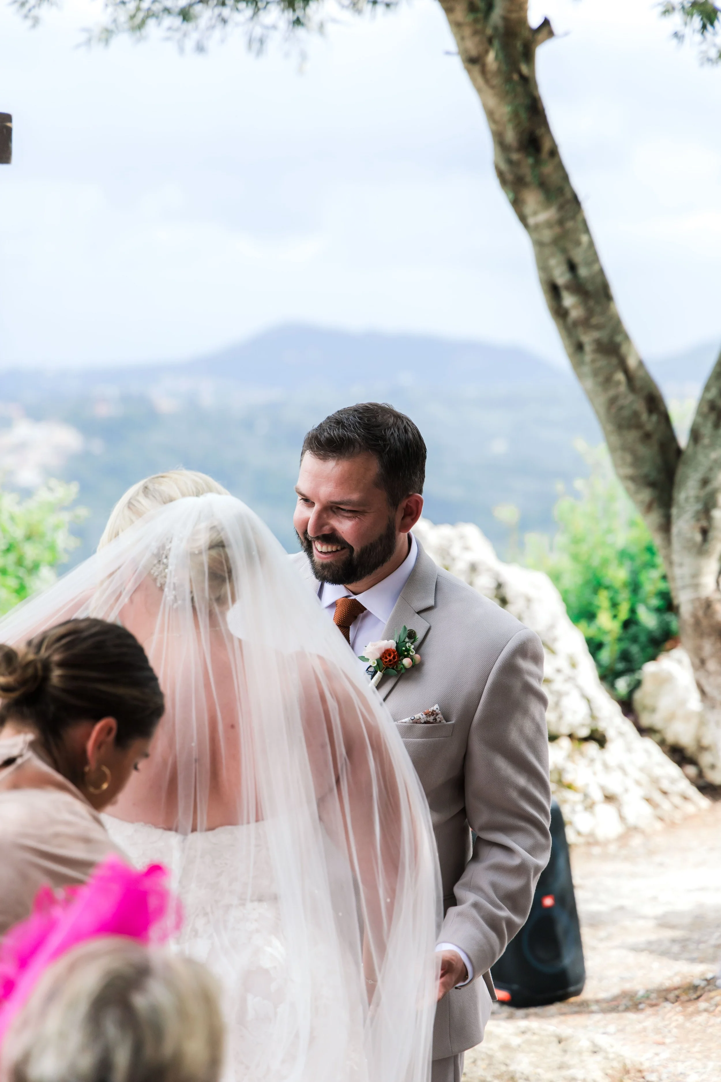 A bride and groom exchanging vows outdoors, with a woman assisting, trees, rocks, and a scenic landscape with mountains in the background.