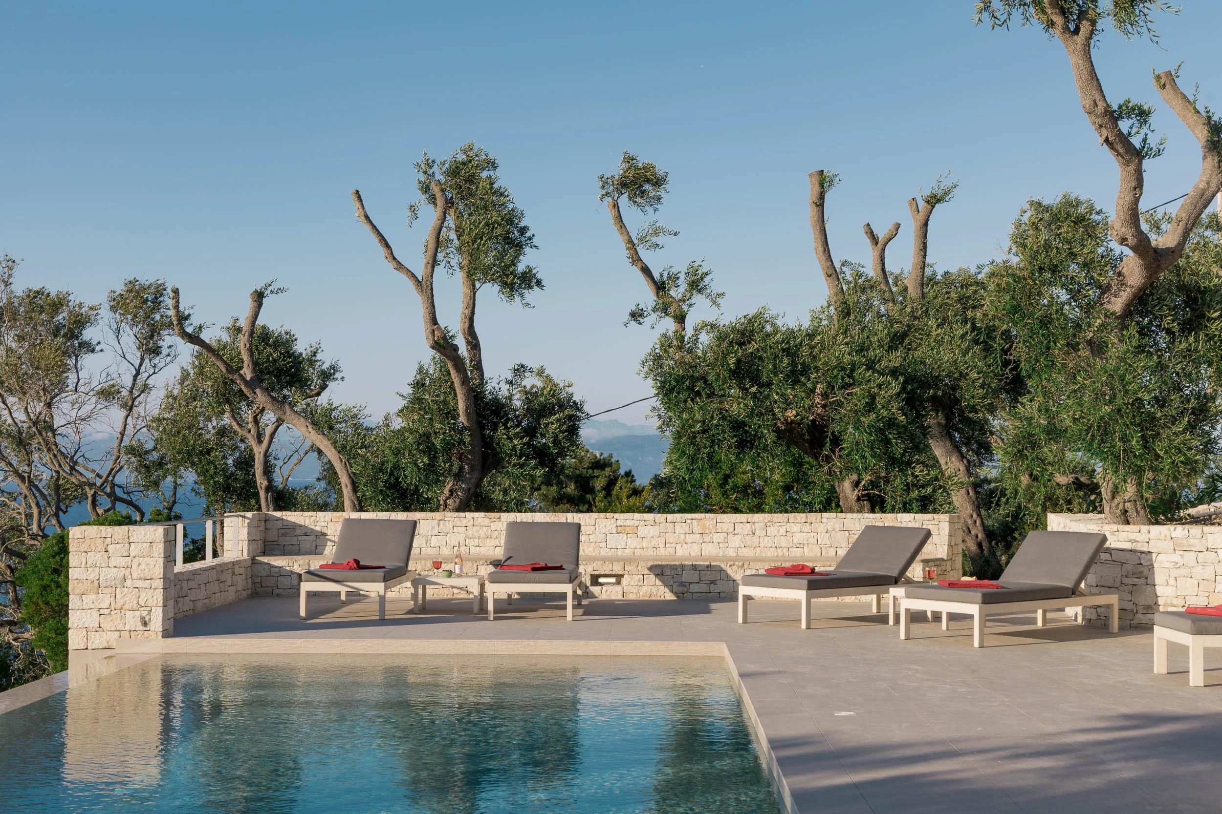 Poolside area with four lounge chairs, small tables, and a stone wall, with trees and a blue sky in the background.