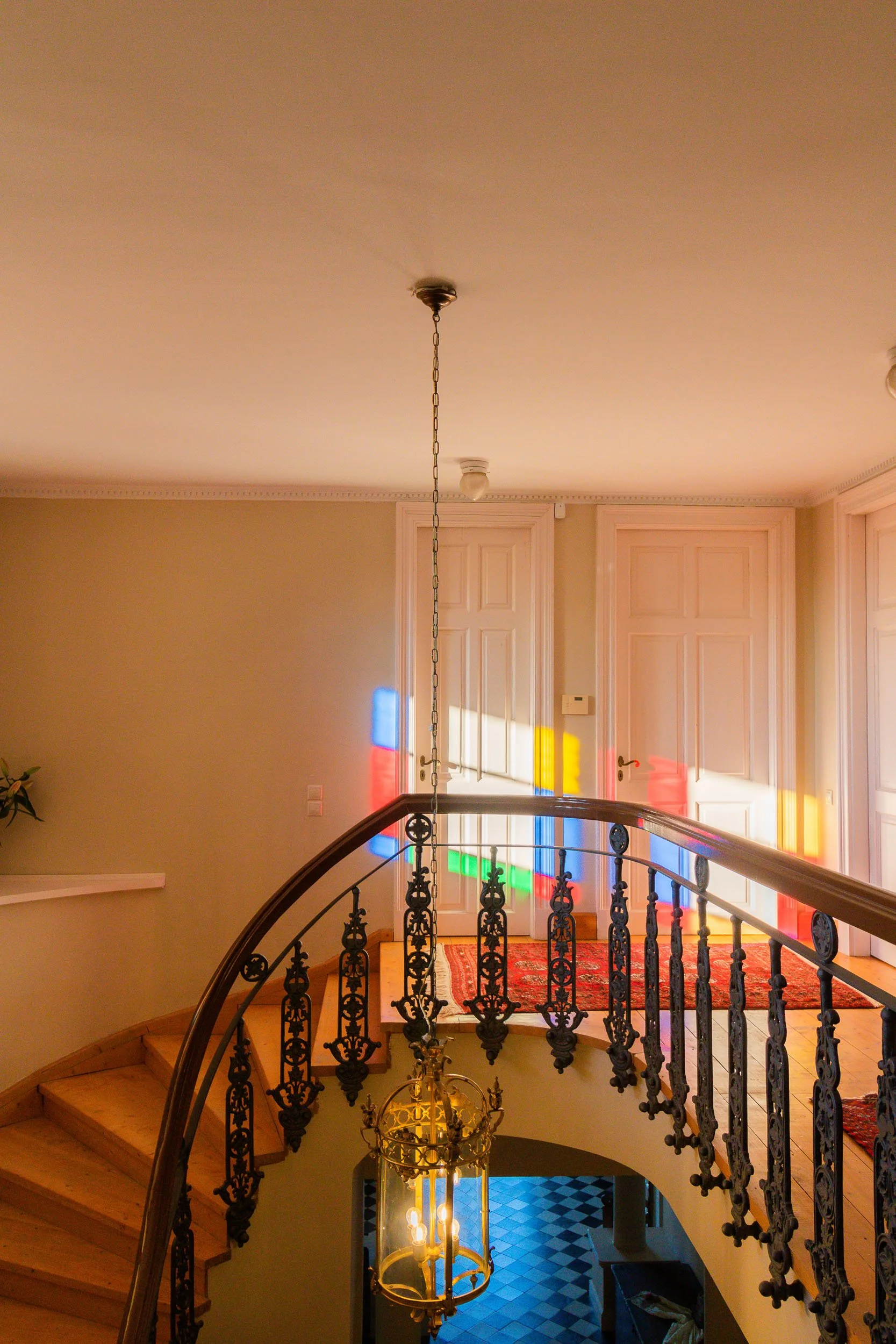 Interior of a house with a curved staircase, iron railing, and stained glass windows casting colorful light on the door and wall.