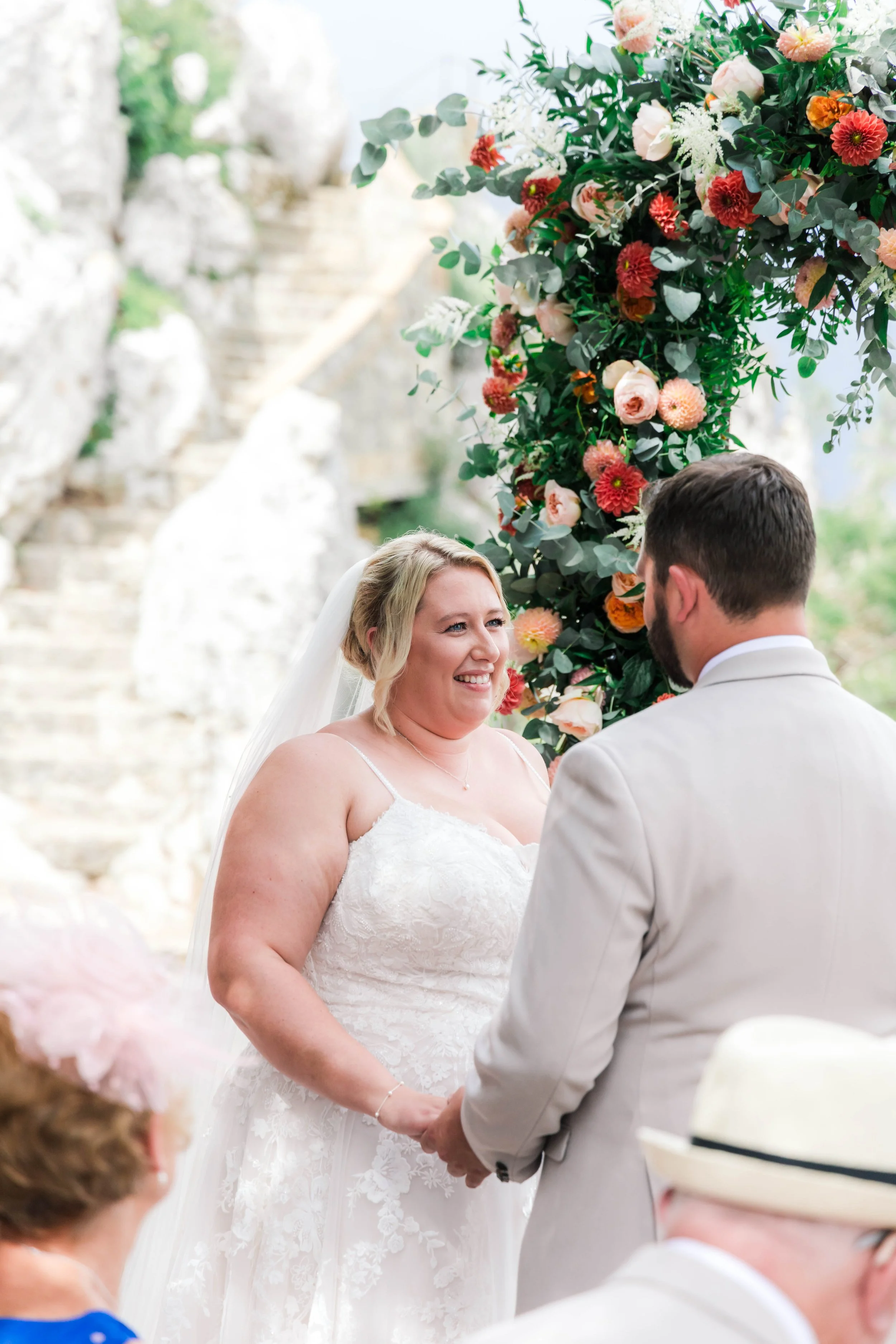 A bride and groom exchanging vows during their wedding ceremony outdoors, with a floral arch and rocky background.