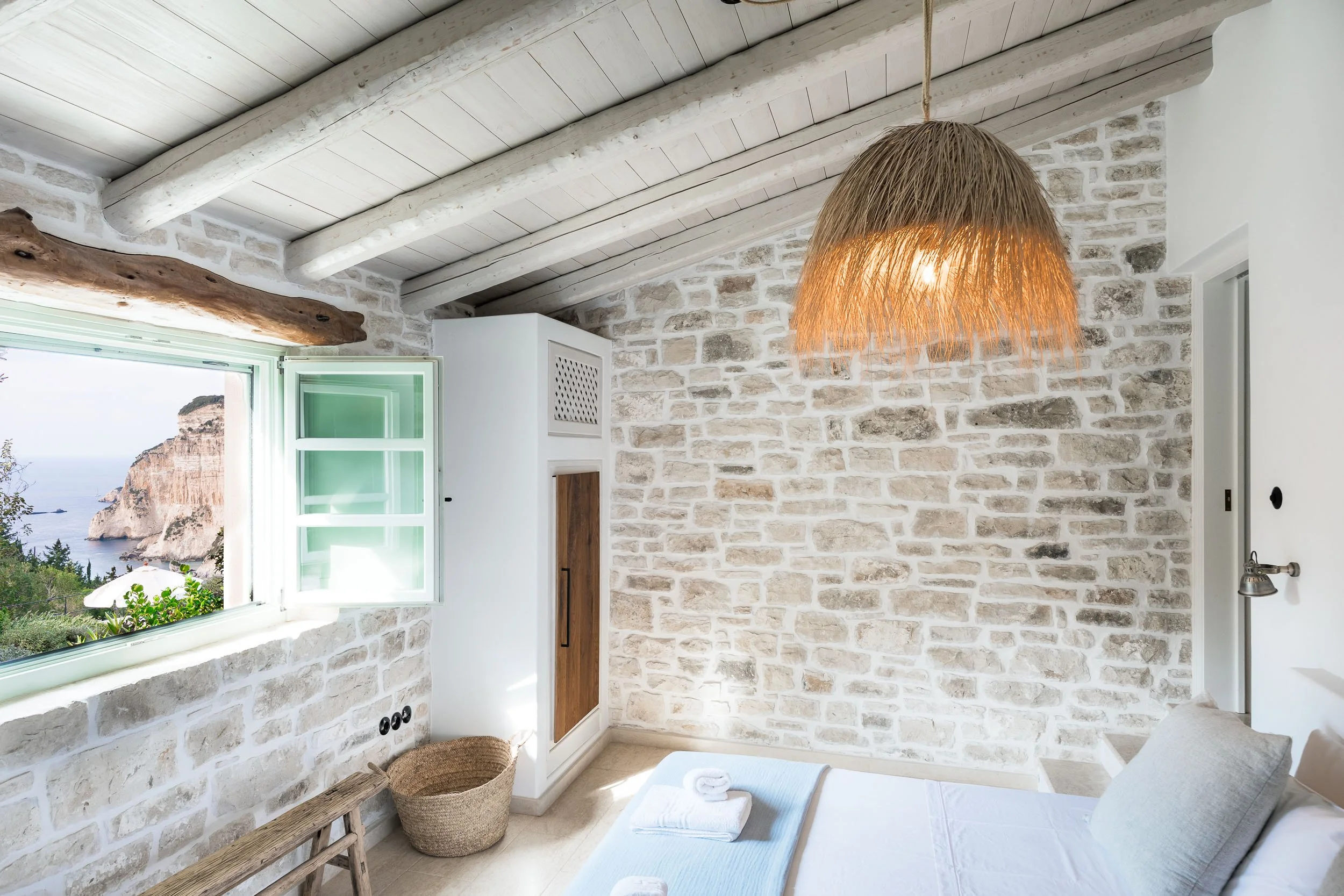 Cozy bedroom with white brick walls, a window with a view of cliffs and ocean, a straw pendant light, and a bed with white linens.