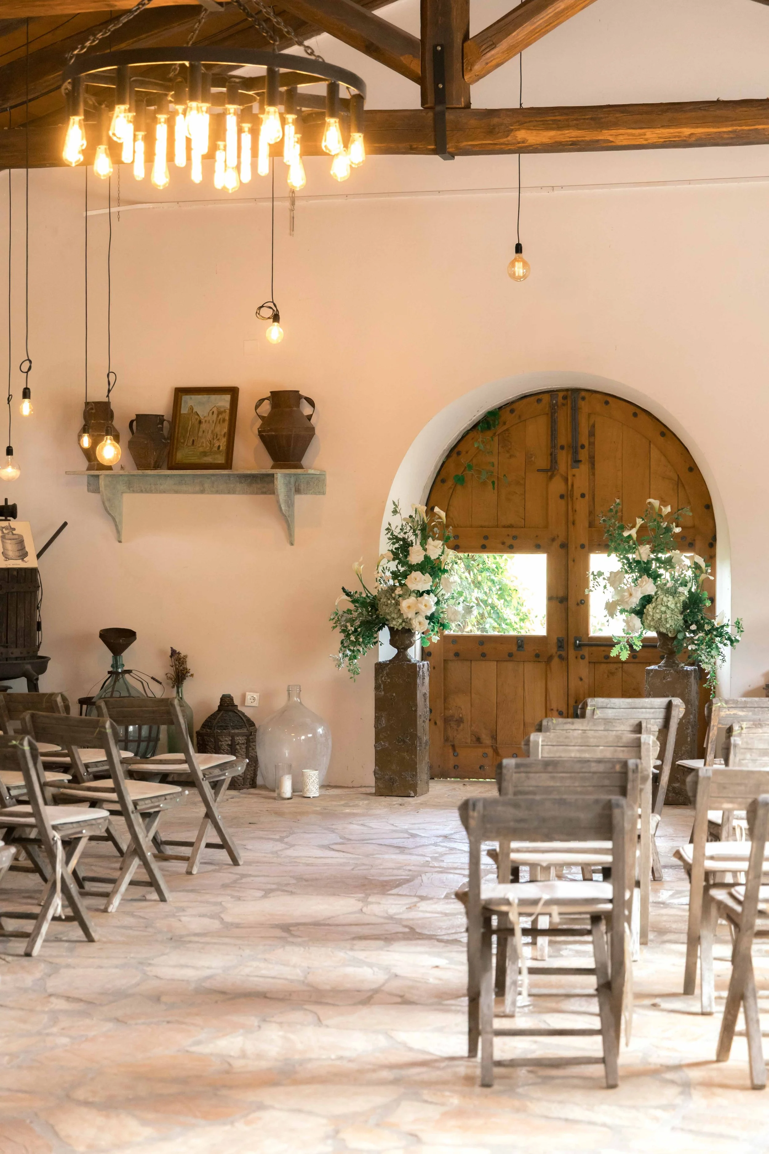 Interior of a rustic wedding or event venue with wooden chairs, large floral arrangements in vases, a wooden door with small windows, hanging light bulbs, and a chandelier with multiple vintage-style bulbs.
