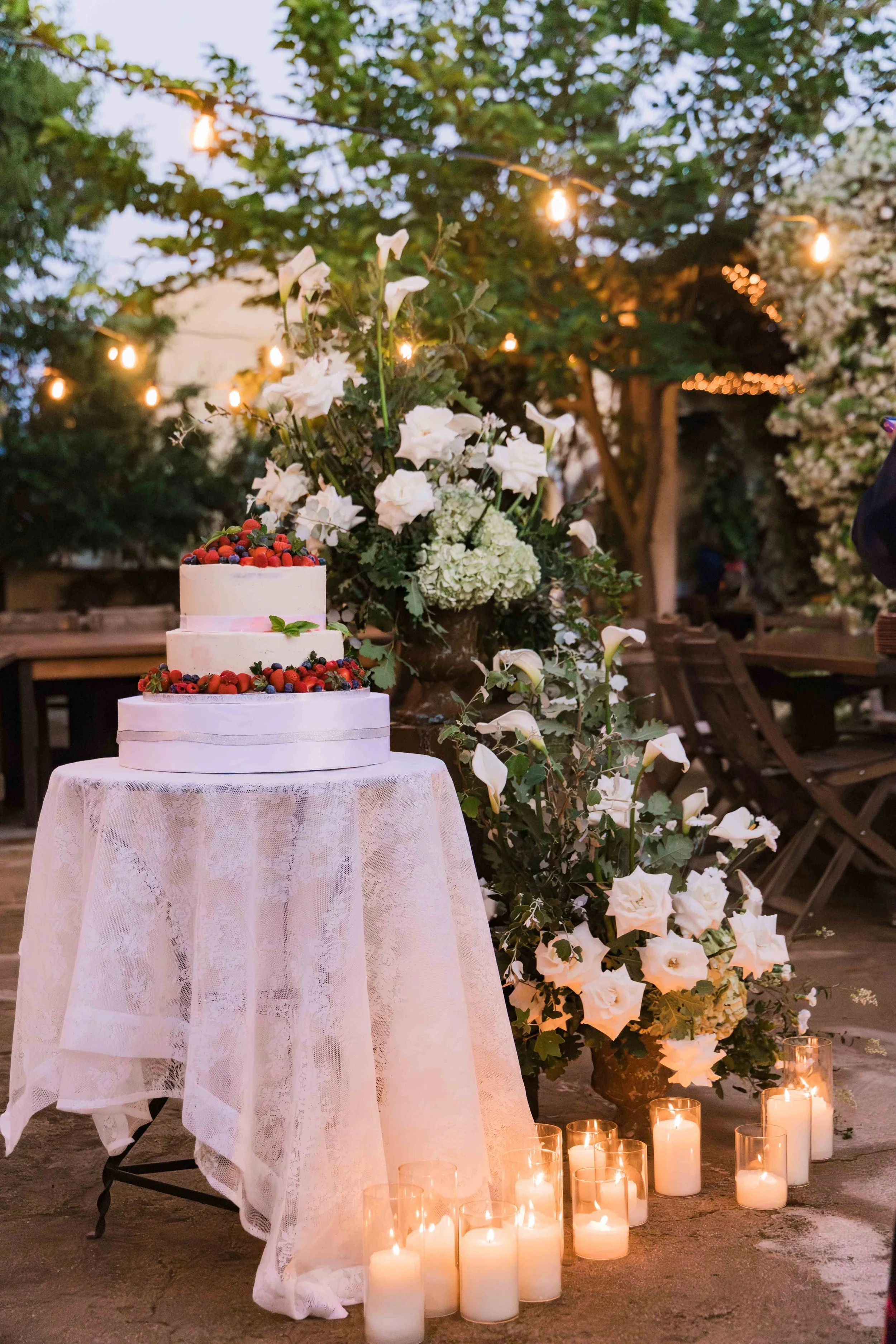 A three-tier white wedding cake decorated with berries, placed on a round table covered with lace, surrounded by white flowers and candles in glass holders, set outdoors with string lights in the background.