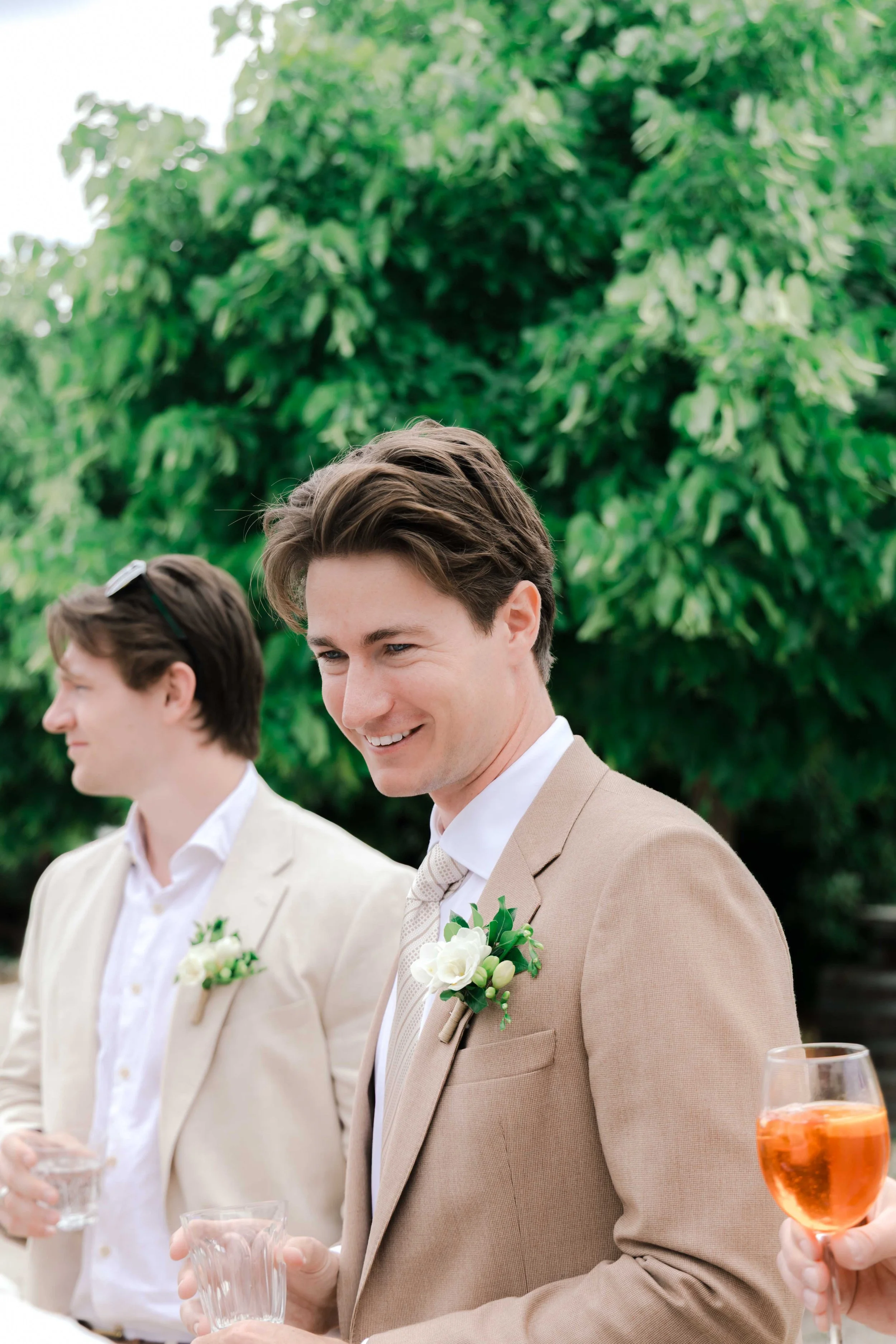 A smiling man in a beige suit with a white shirt and tie, holding a glass, standing outdoors with a lush green tree in the background. Another man in a light suit and boutonniere is partially visible beside him, also holding a glass.