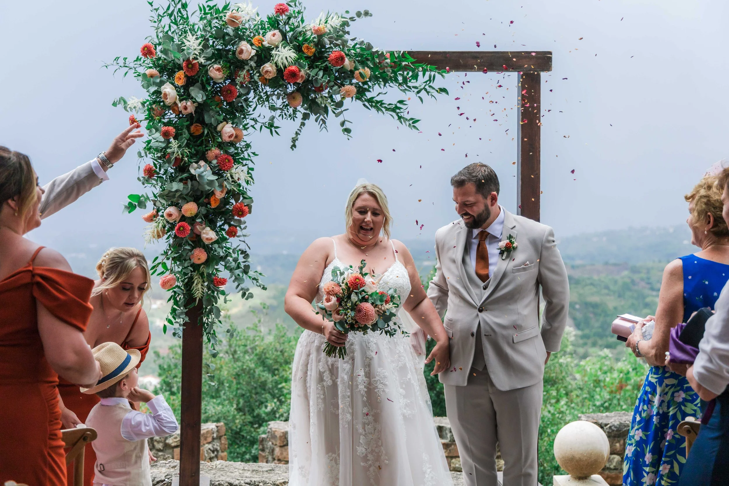 A bride and groom at their wedding ceremony, holding hands and smiling under a floral arch. The bride is in a white dress, holding a bouquet, and the groom is in a light suit. Guests are celebrating around them, with some throwing flower petals.