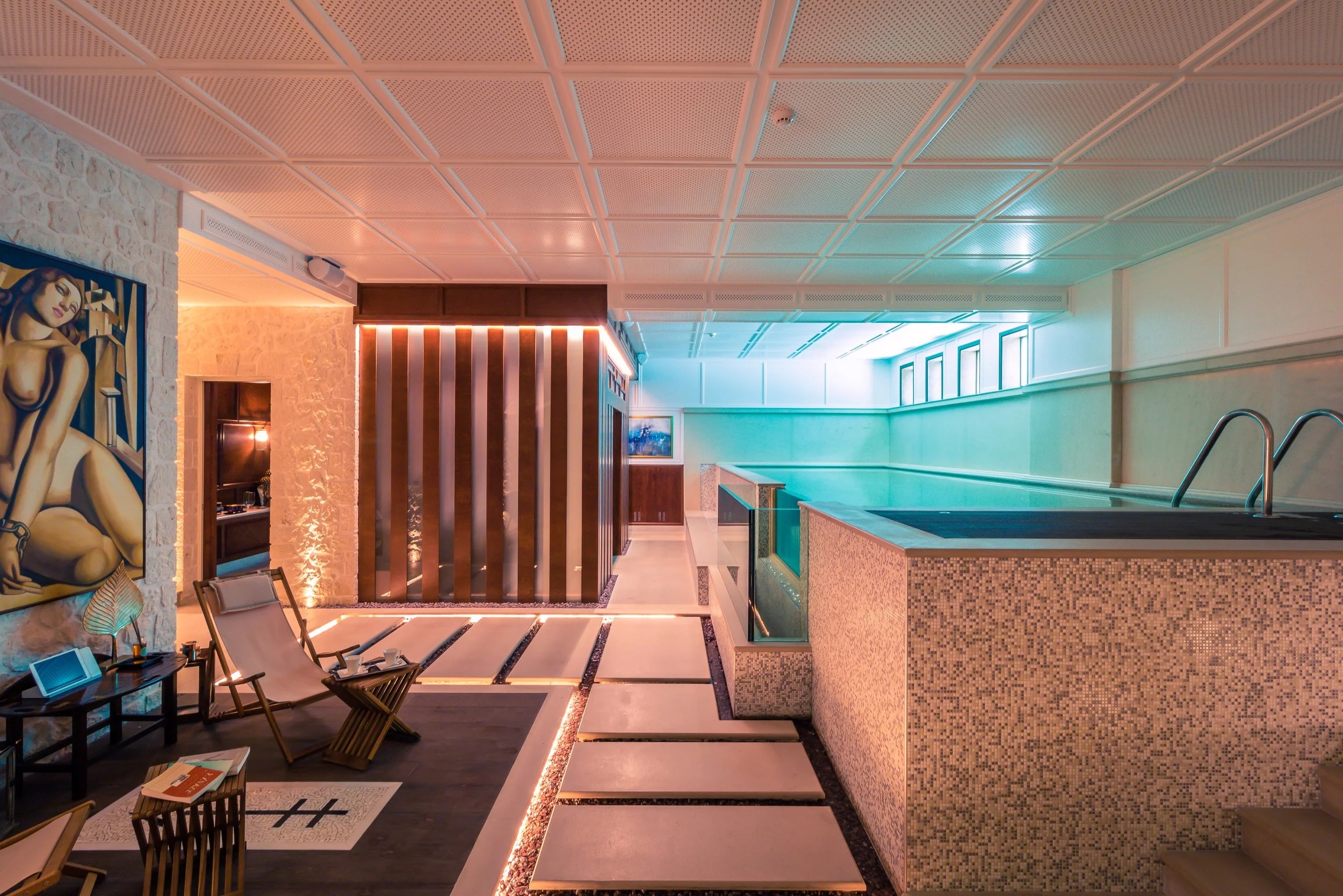 Indoor luxury spa with a swimming pool, seating area with lounge chairs, modern decor, artwork on walls, ambient lighting, and step access to the pool.