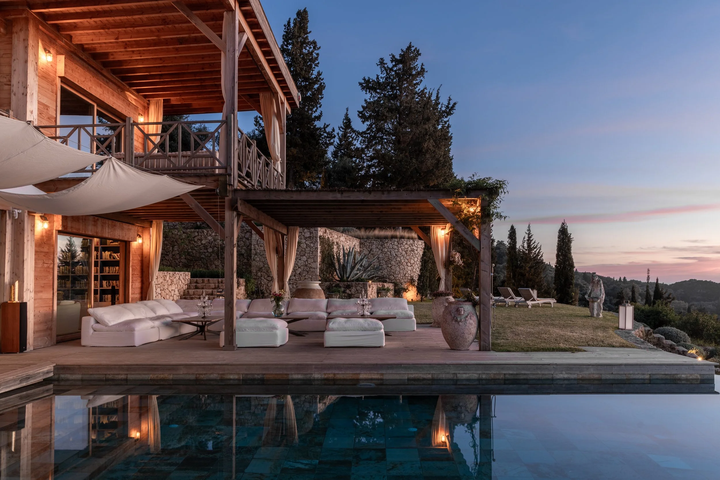Luxury house with outdoor patio, pool, white sectional sofa, large vases, string lights, and lounge chairs under a sunset sky, surrounded by trees and landscape.