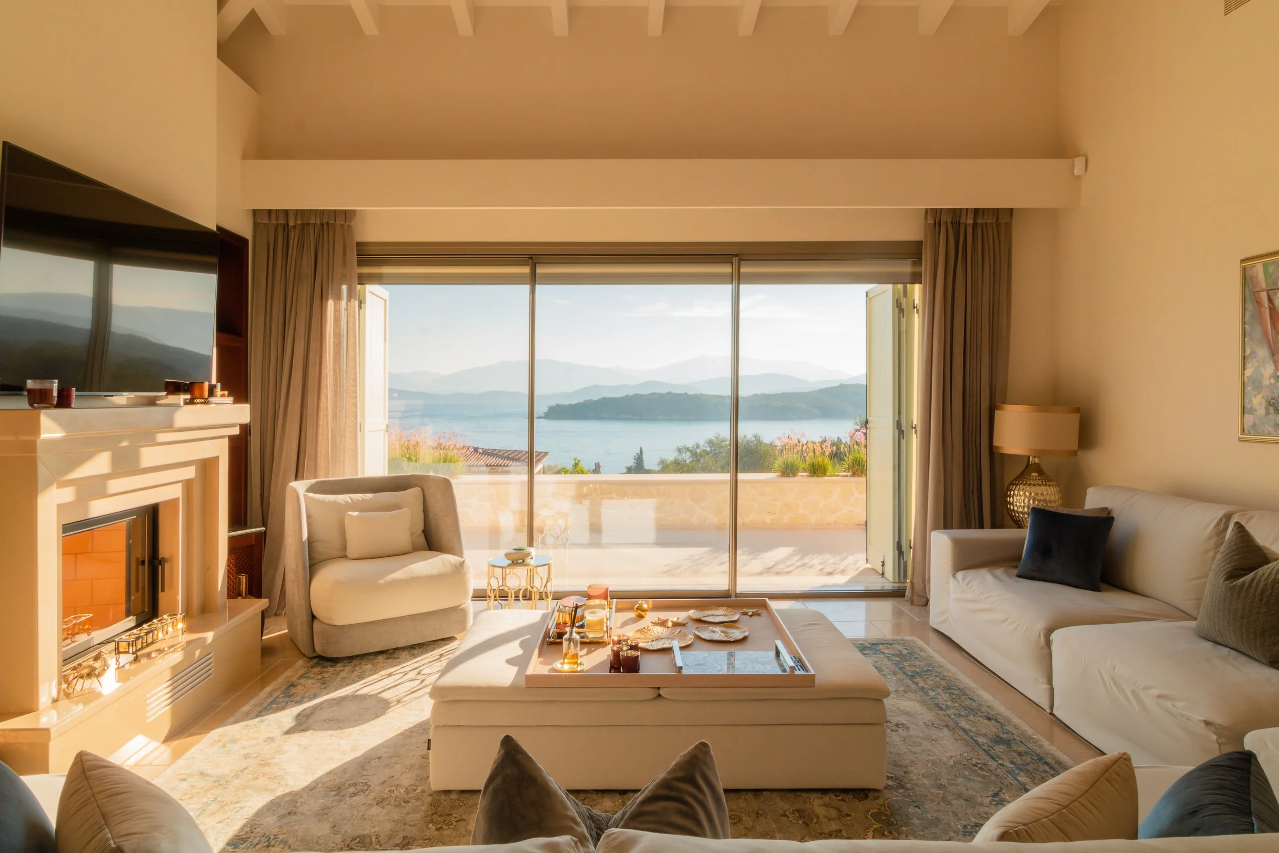 Living room with large sliding glass doors opening to a balcony with a view of a lake and mountains in the background, beige sofas, a white armchair, a fireplace, and a tray with breakfast items on a ottoman.