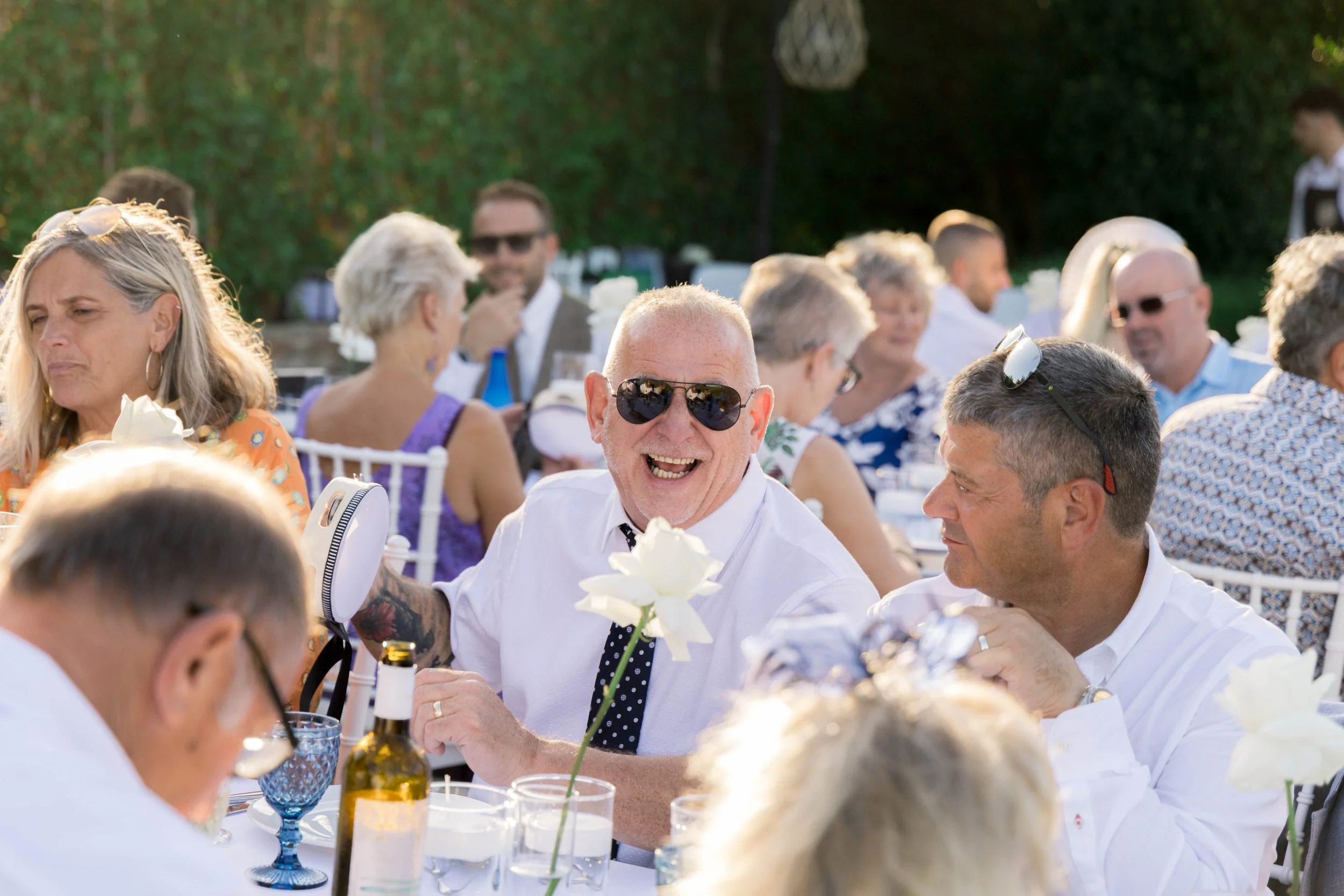 Group of people at an outdoor celebration or party, sitting at a table with floral decorations, drinks, and food, smiling and interacting with each other.