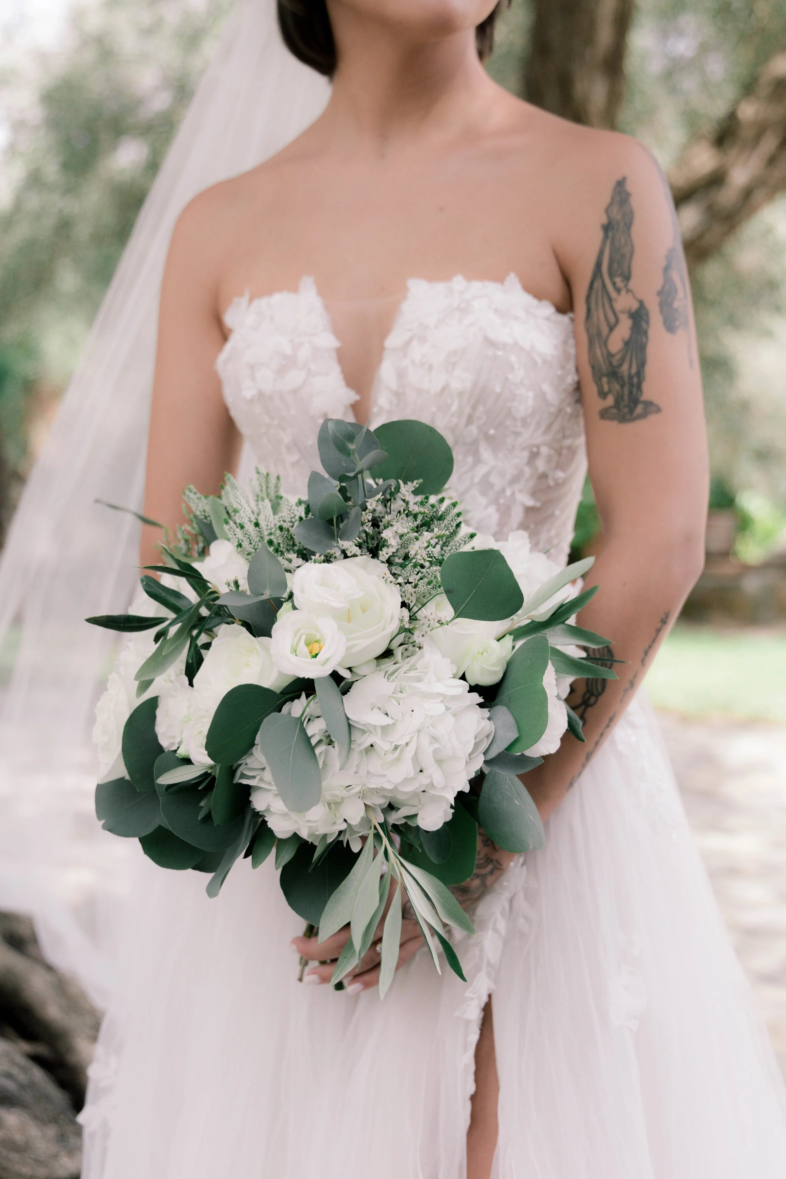Bride holding a bouquet of white flowers and green leaves, wearing a strapless wedding dress with lace details, outdoors on a sunny day.