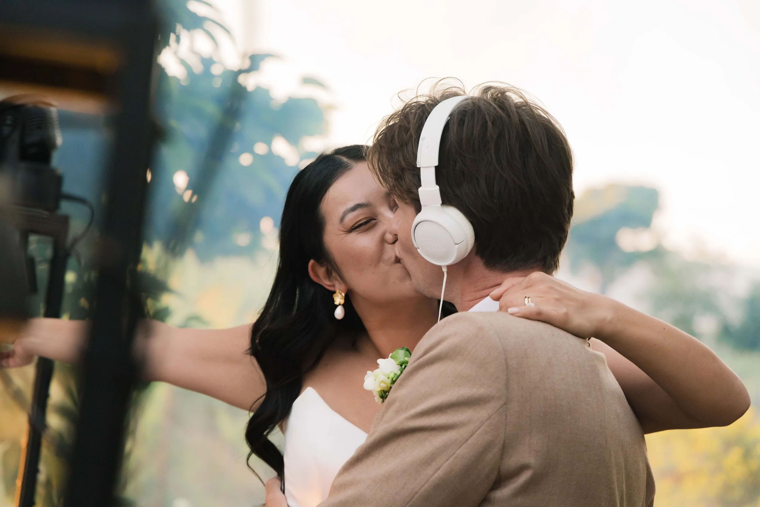 A woman and a man share a kiss outdoors, with the woman wearing a white dress and pearl earrings, and the man wearing a tan suit and headphones, with greenery and trees in the background.