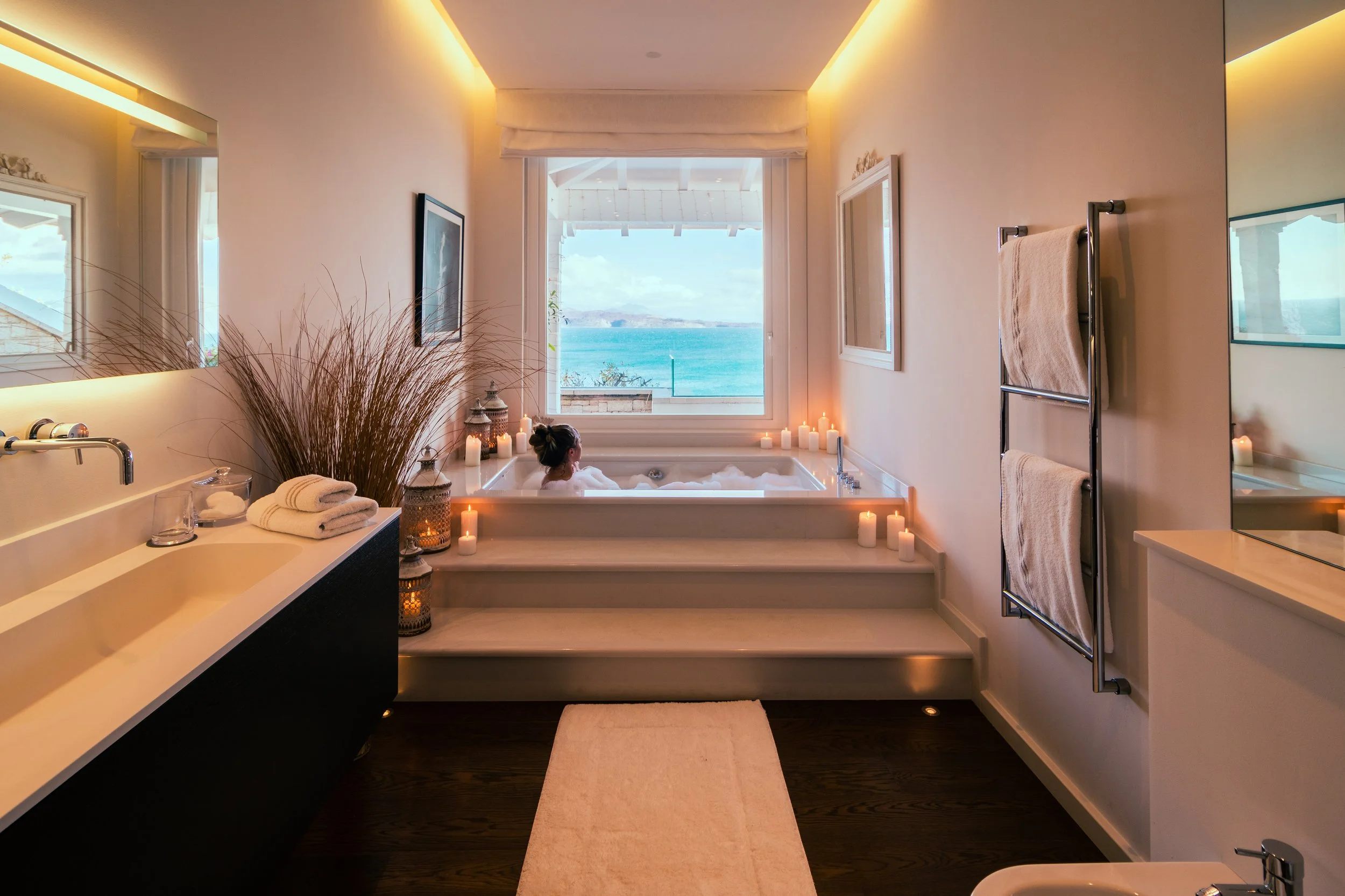 A woman relaxing in a bathtub with a view of the ocean through a large window, candles surrounding the tub, and a bathroom with towels and a sink.