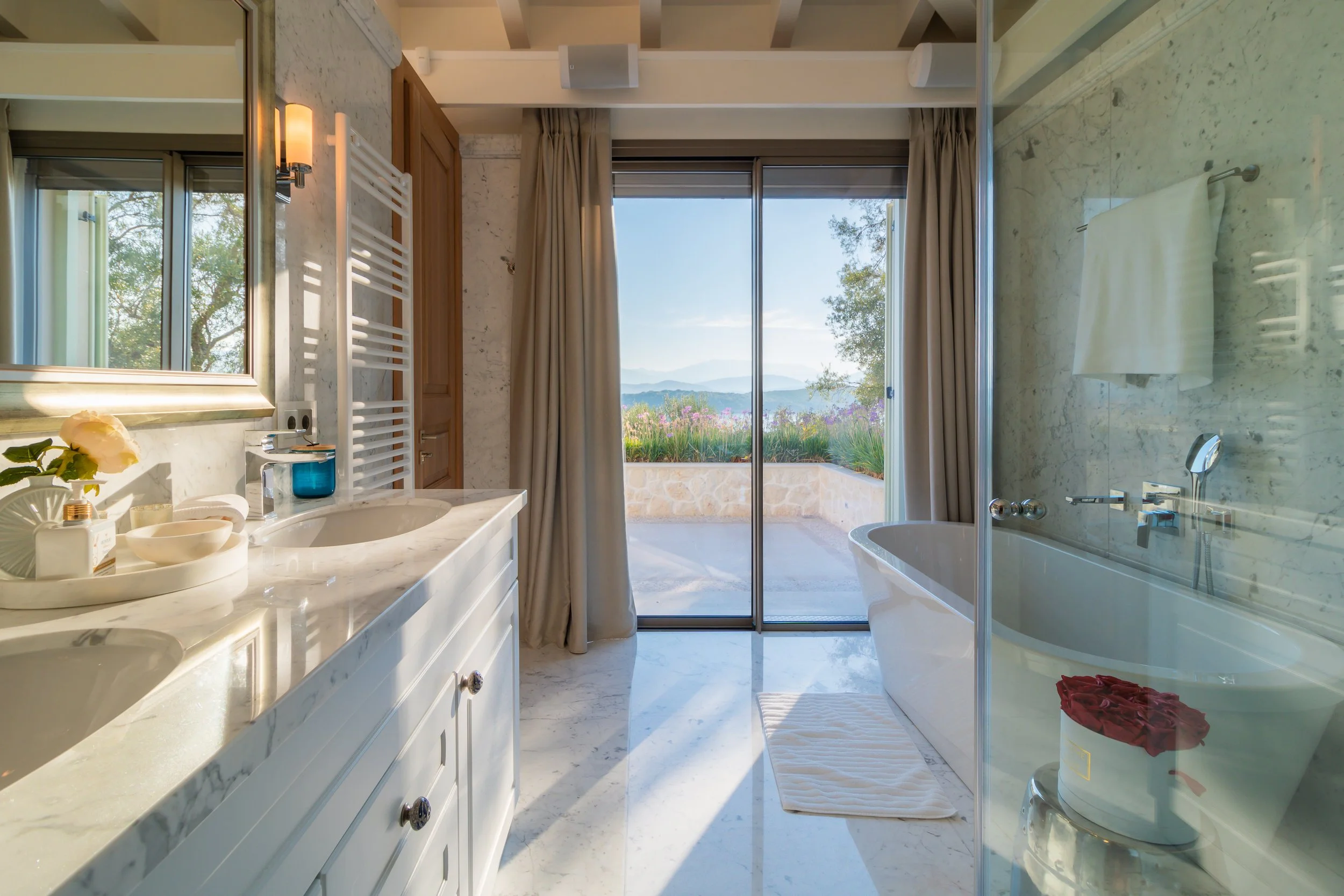 Bathroom with double vanity, bathtub, shower, and large glass sliding door leading to a balcony with a view of mountains and greenery.