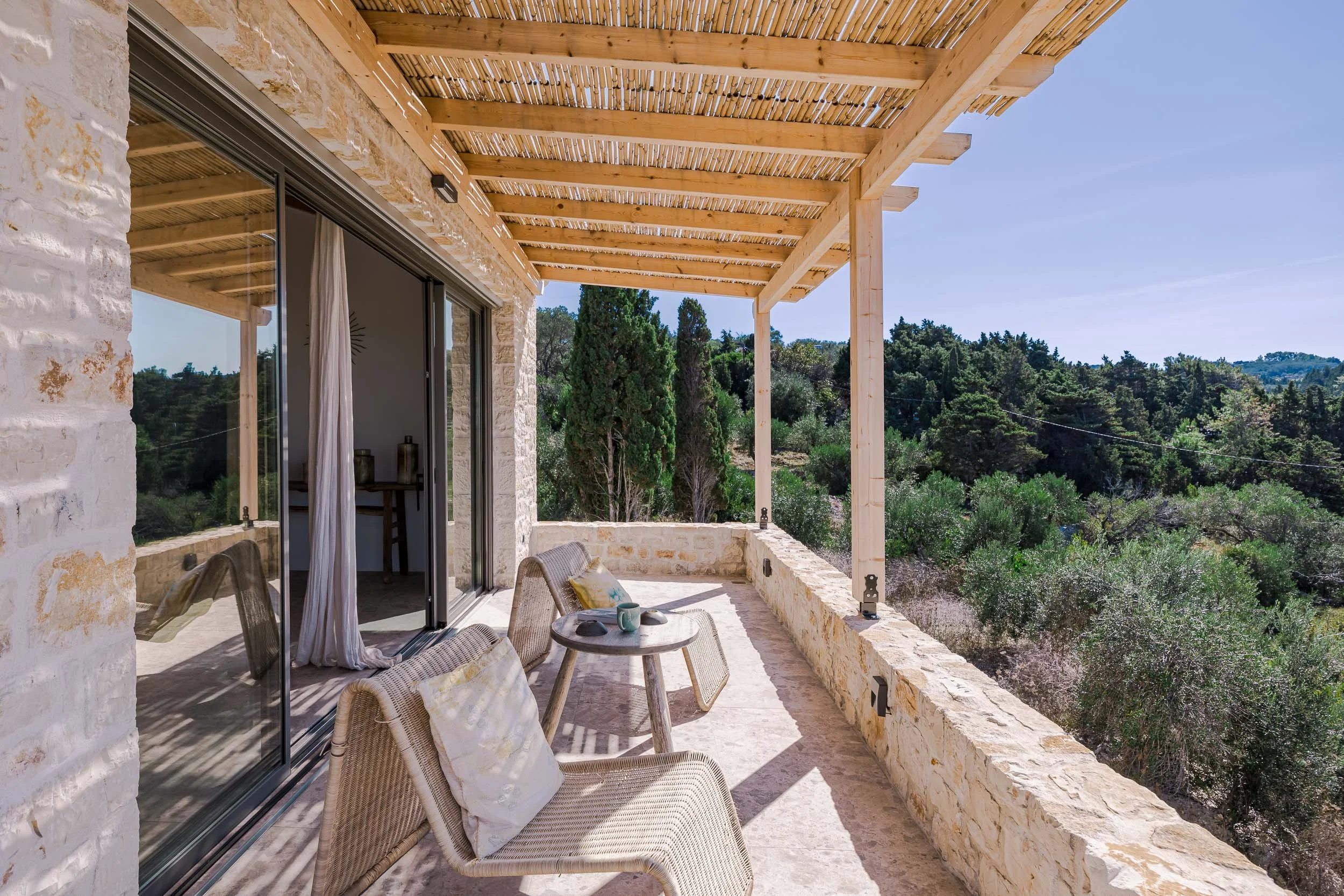 A spacious outdoor balcony with stone walls, wooden beams, and a view of green trees and hills, featuring two wicker chairs with cushions and a small round table.