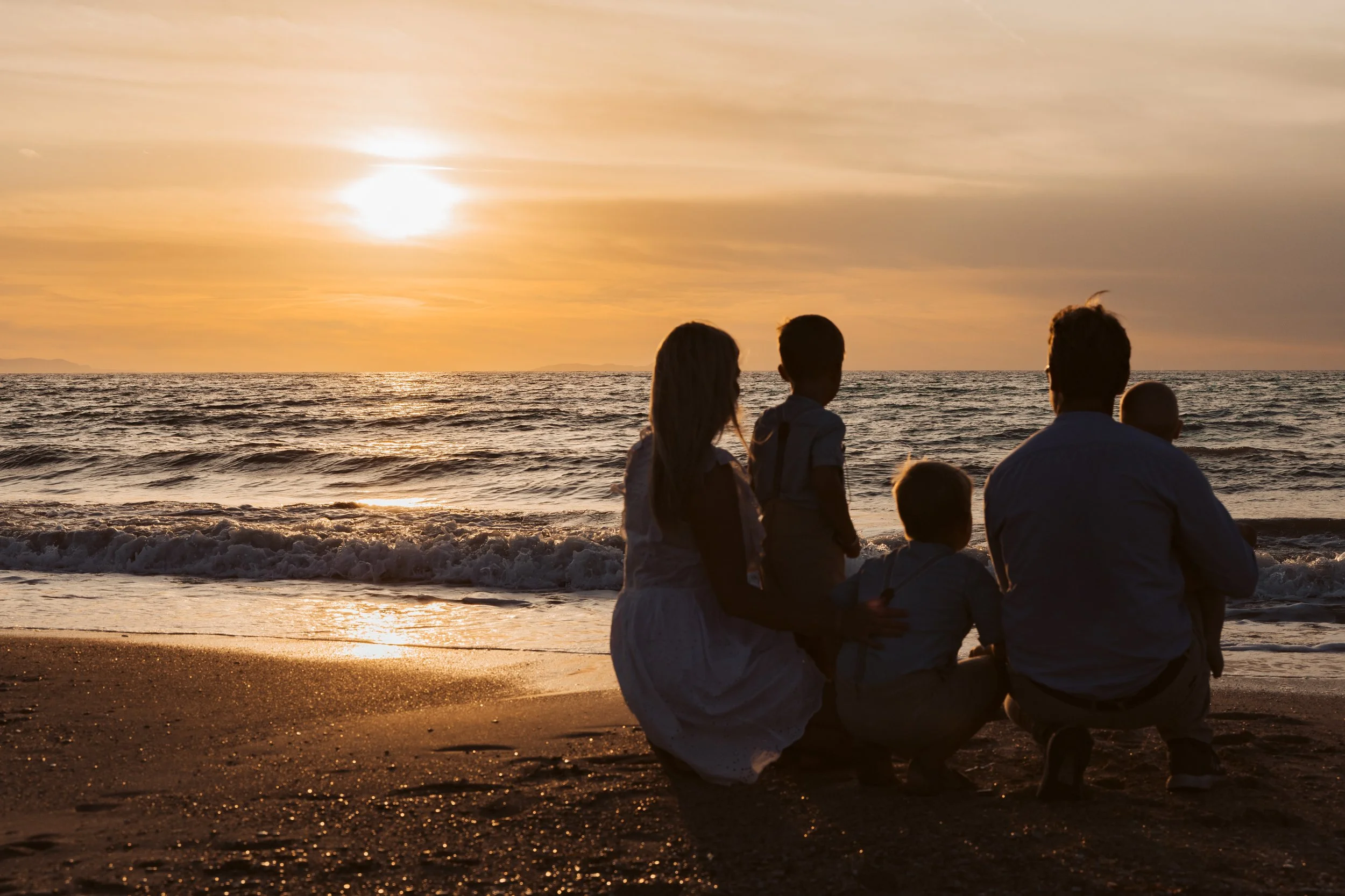 Group of five people, including children and adults, sitting on a sandy beach during sunset, looking out at the ocean waves.