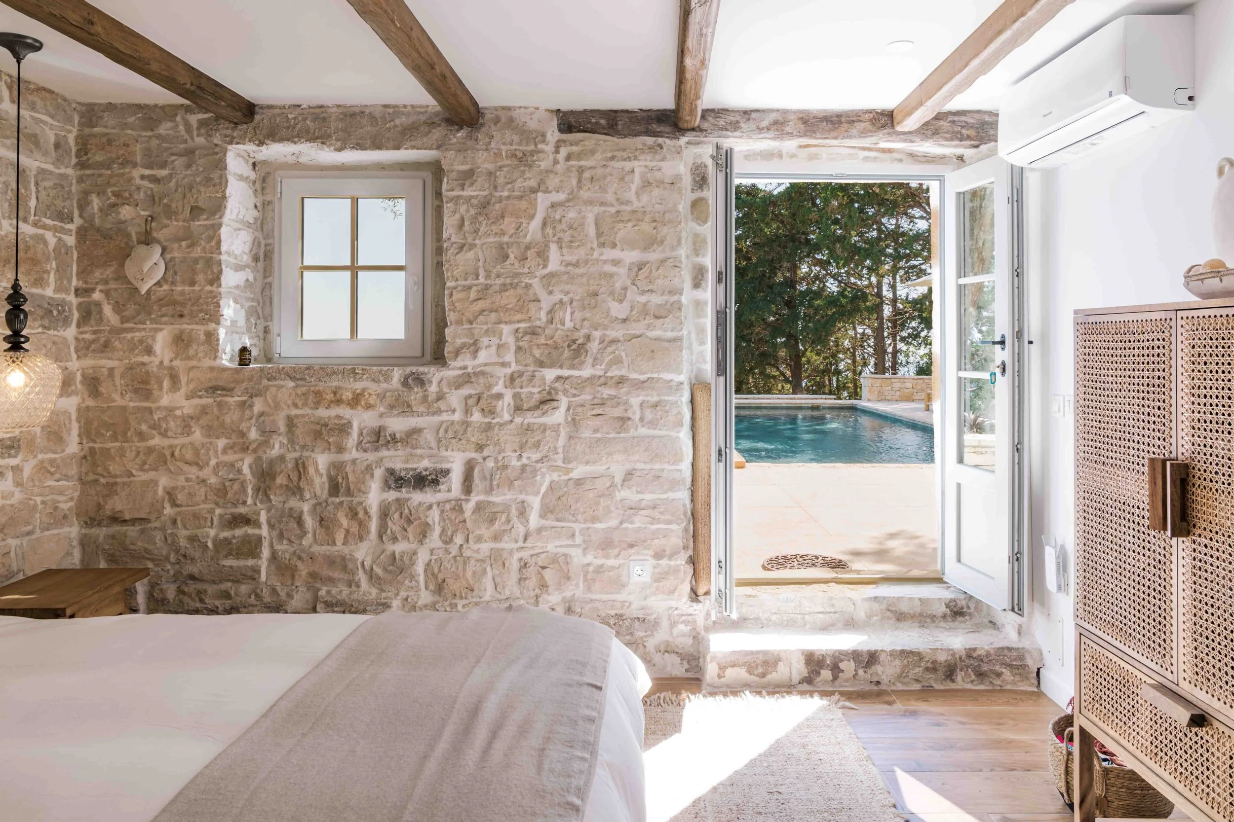 Interior of a bedroom with stone wall, small window, and open door leading to an outdoor pool and patio area with trees