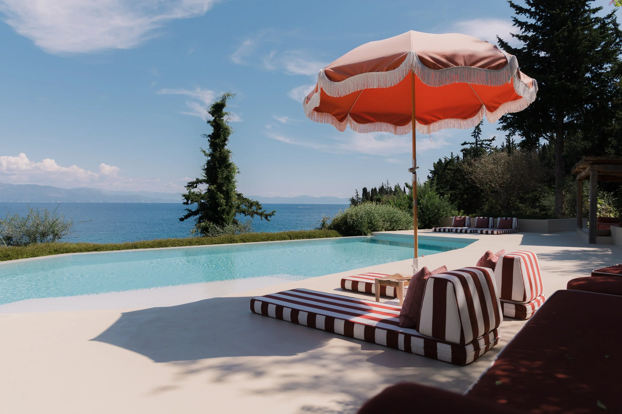 A swimming pool with striped lounge chairs and a large peach-colored beach umbrella overlooking a lake with distant mountains and trees.
