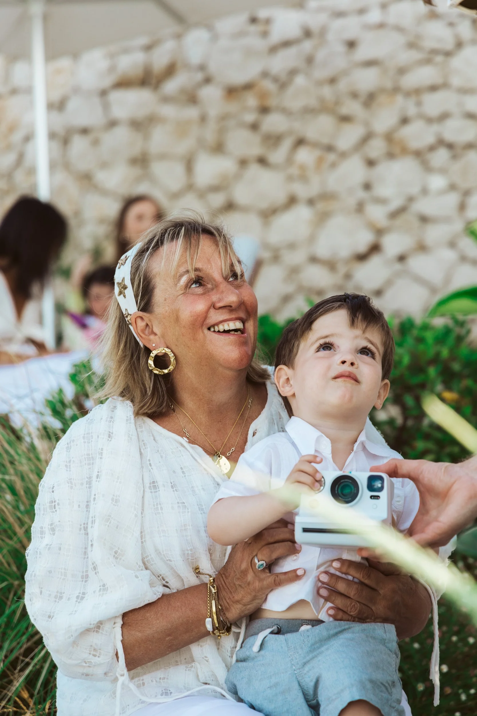A smiling older woman with gold hoop earrings, wearing a white blouse with gold jewelry, holds a young boy in her arms, who is looking up while holding a small instant camera. They are outdoors in front of a stone wall, with other people visible in t