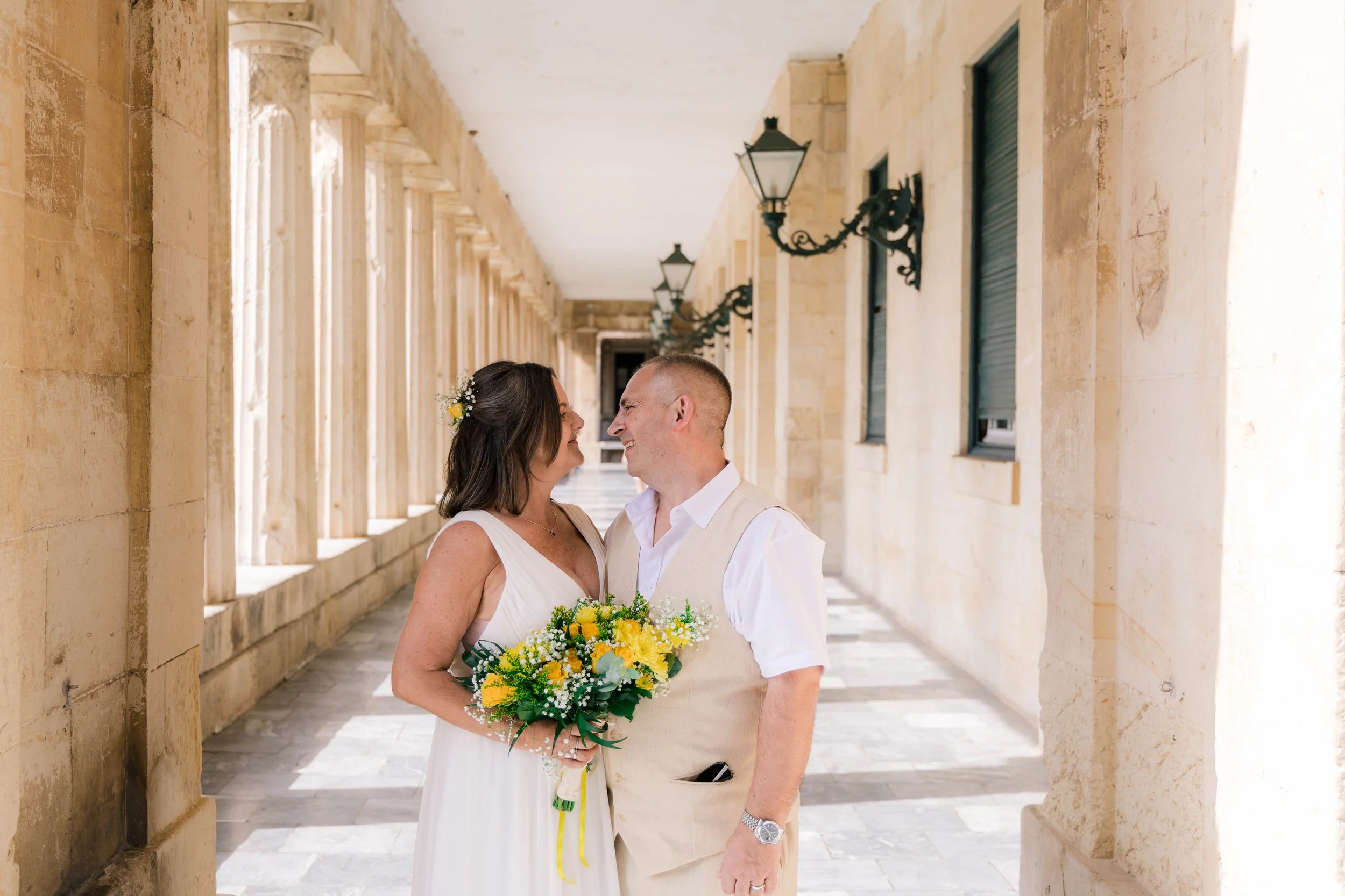 A couple in wedding attire standing close and smiling at each other under a colonnade with stone walls and black lamp posts.