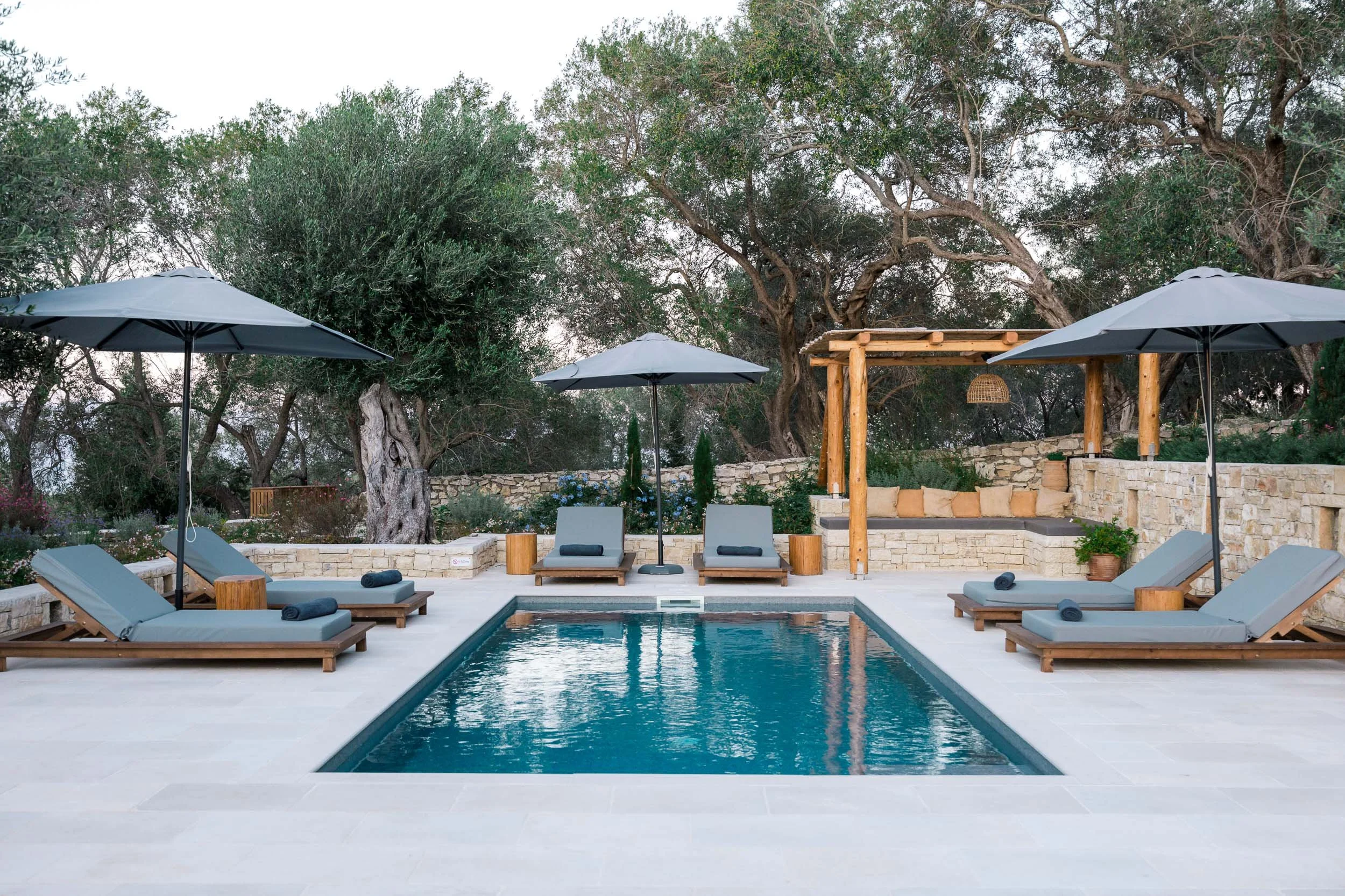 Outdoor swimming pool with six loungers, umbrellas, and a shaded seating area surrounded by trees and stone walls.