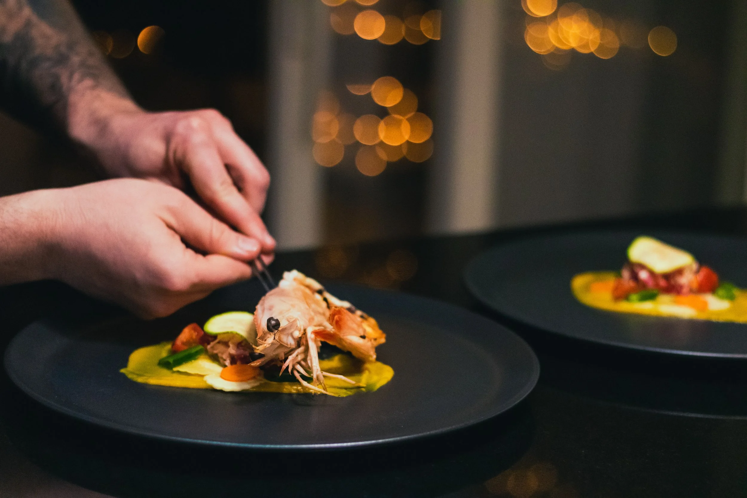 A chef is garnishing two black plates of gourmet seafood dishes with a shrimp and colorful vegetables, with warm bokeh lights in the background.