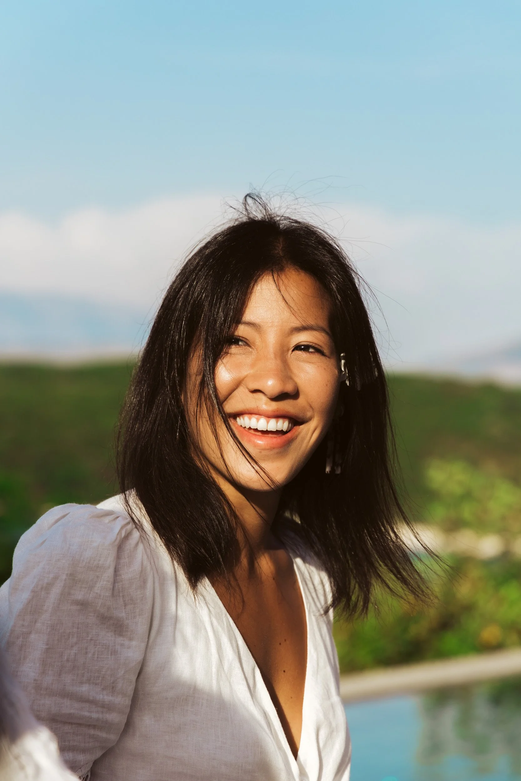 A woman with shoulder-length black hair smiling outdoors against a cloudy sky and green landscape.