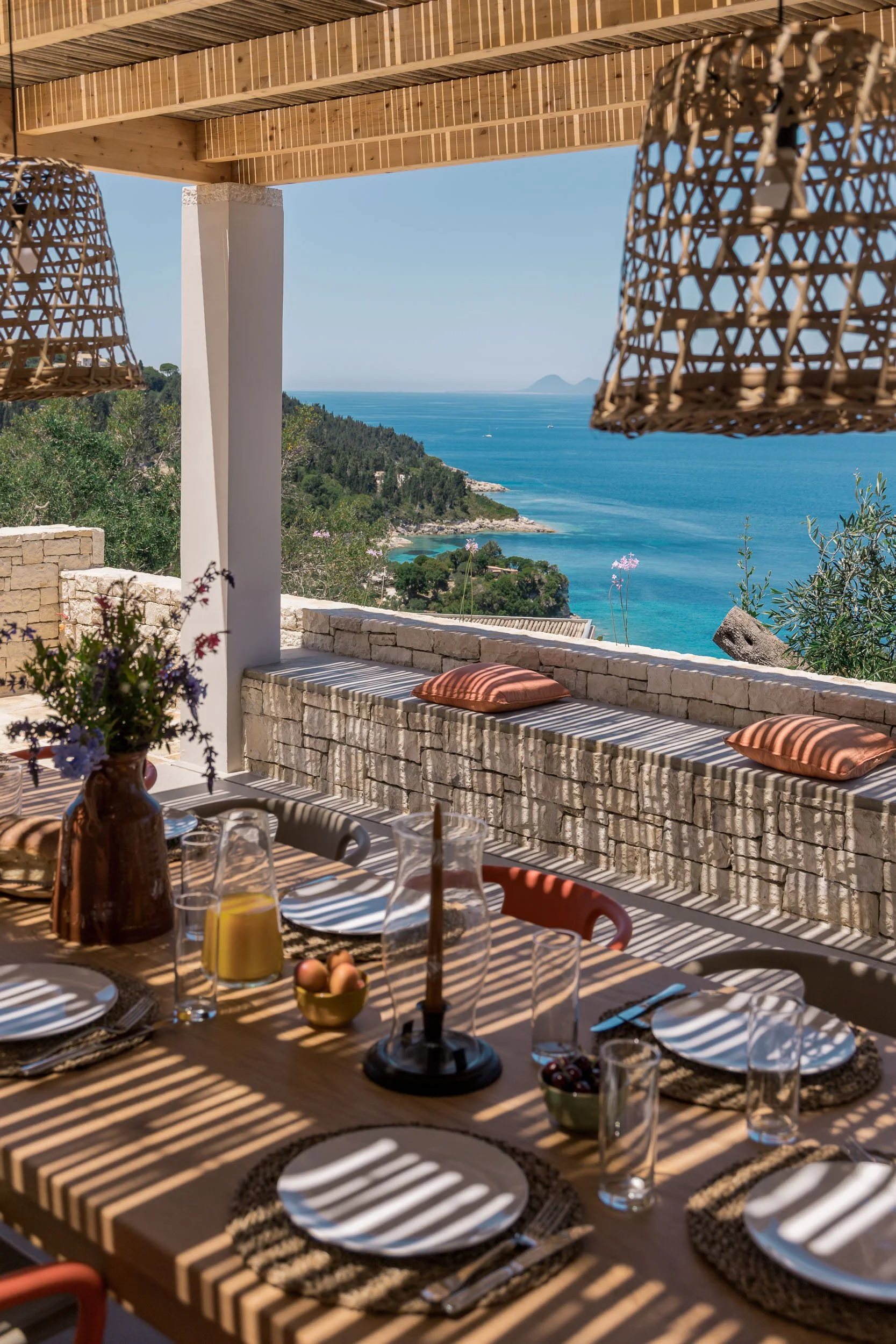 Dining table set for a meal on a balcony with a view of the ocean, rocky coastline, and distant islands, featuring plates, glasses, a pitcher, and a vase of flowers, with wicker lanterns hanging above.
