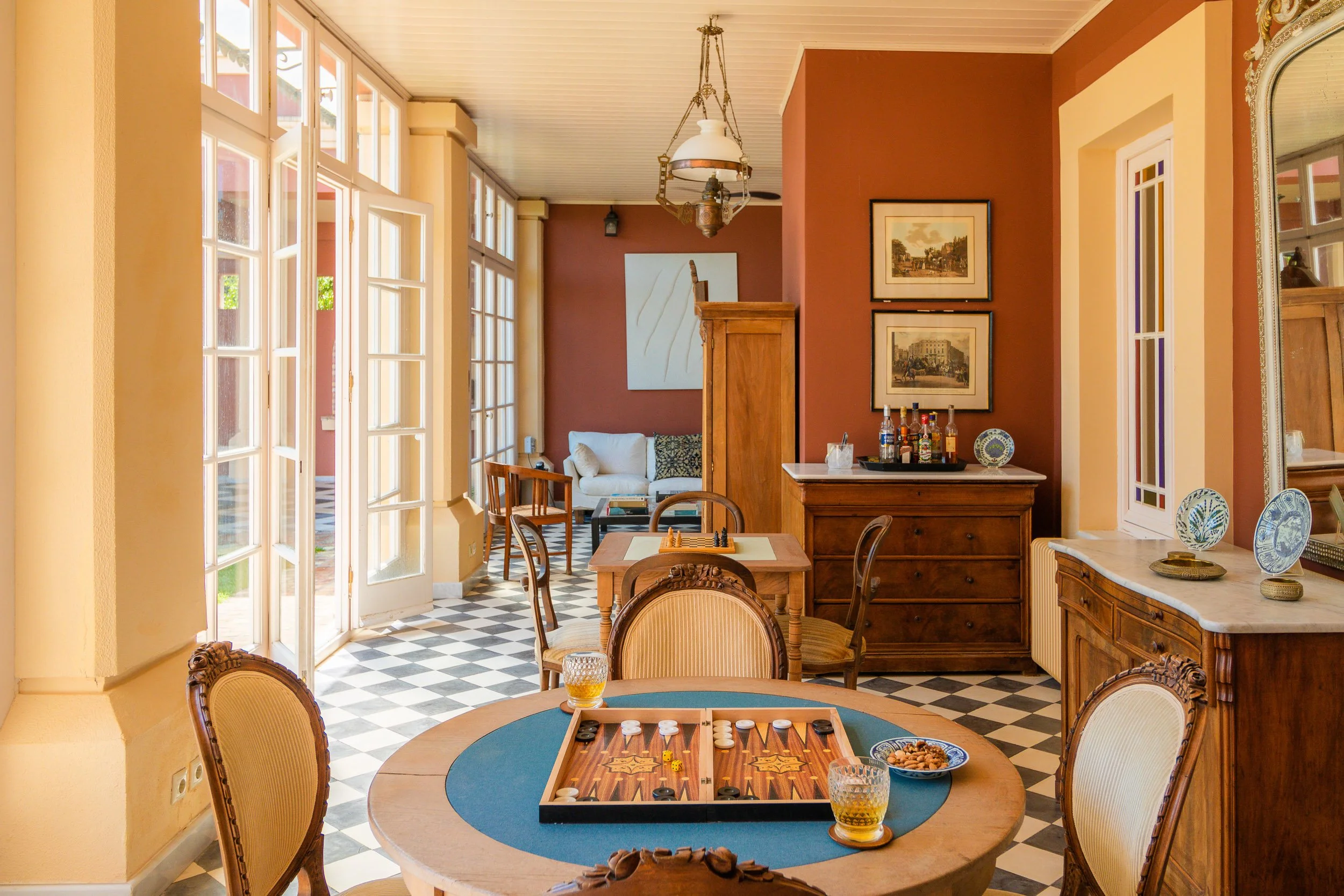 Living room with checkered black and white floor, wooden furniture, a round table with a backgammon game and snacks, a bar with alcohol bottles, white sofa, framed pictures on the wall, windows opening to the outside, and decorative plates.