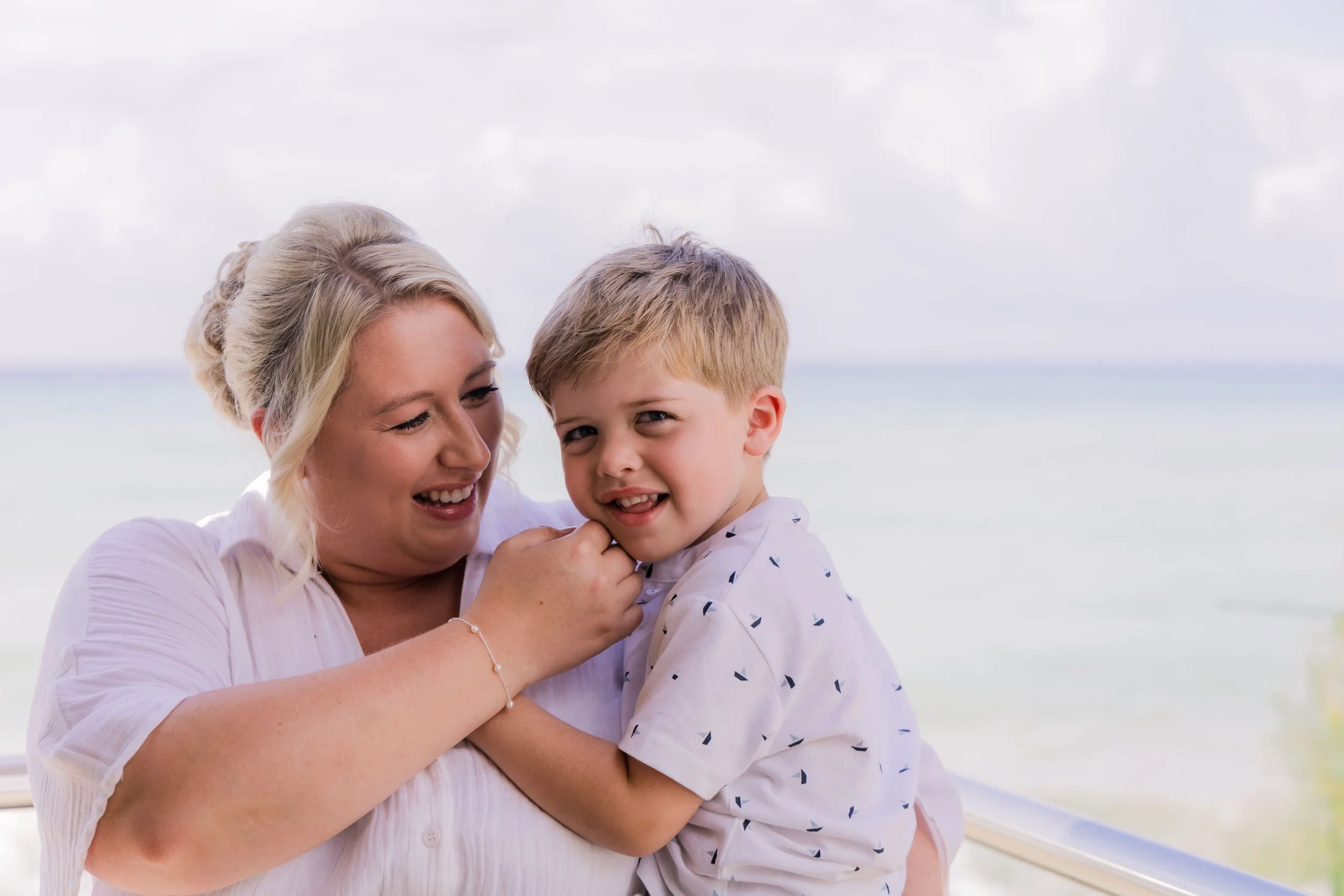 A woman and a young boy laughing and hugging by a window overlooking the beach.