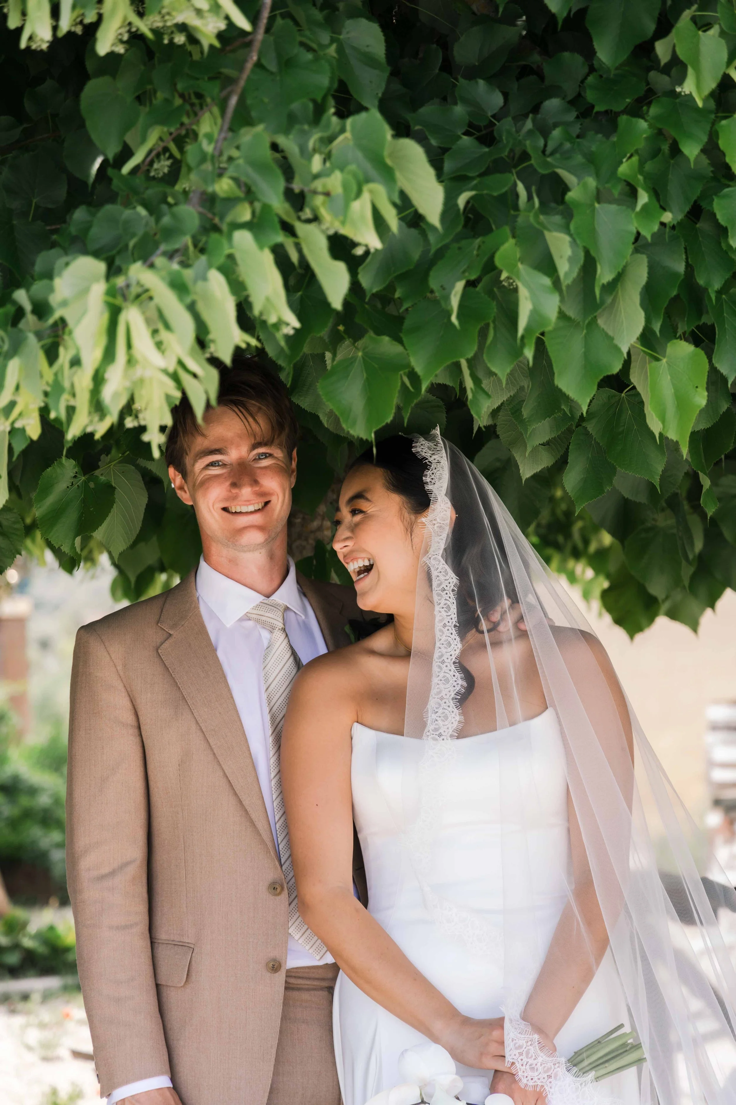 A happy bride and groom standing under a leafy tree, smiling and laughing outdoors on their wedding day. The bride wears a white strapless wedding gown and veil, holding a bouquet of calla lilies. The groom wears a tan suit with a white shirt and pat