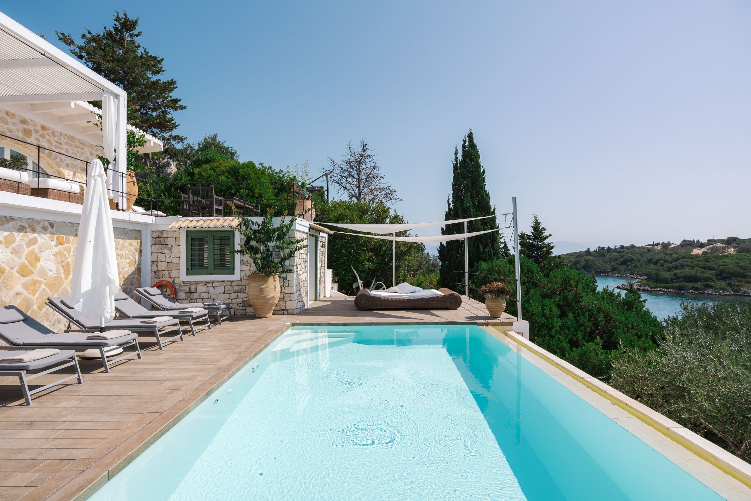 Luxury outdoor swimming pool area with lounge chairs, umbrellas, and scenic view of the ocean and lush greenery.