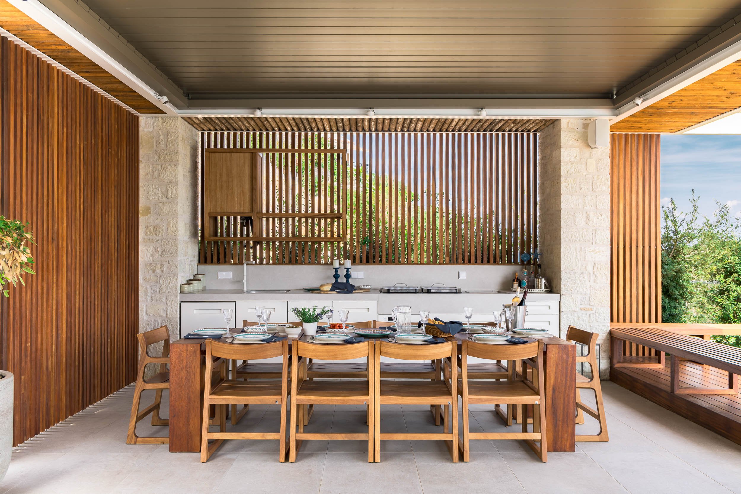 Outdoor dining area with a wooden table set with plates, glasses, and a plant centerpiece, surrounded by wooden chairs; modern kitchen with white cabinets and stone walls in the background; wooden slats on the walls and outdoor greenery visible throu